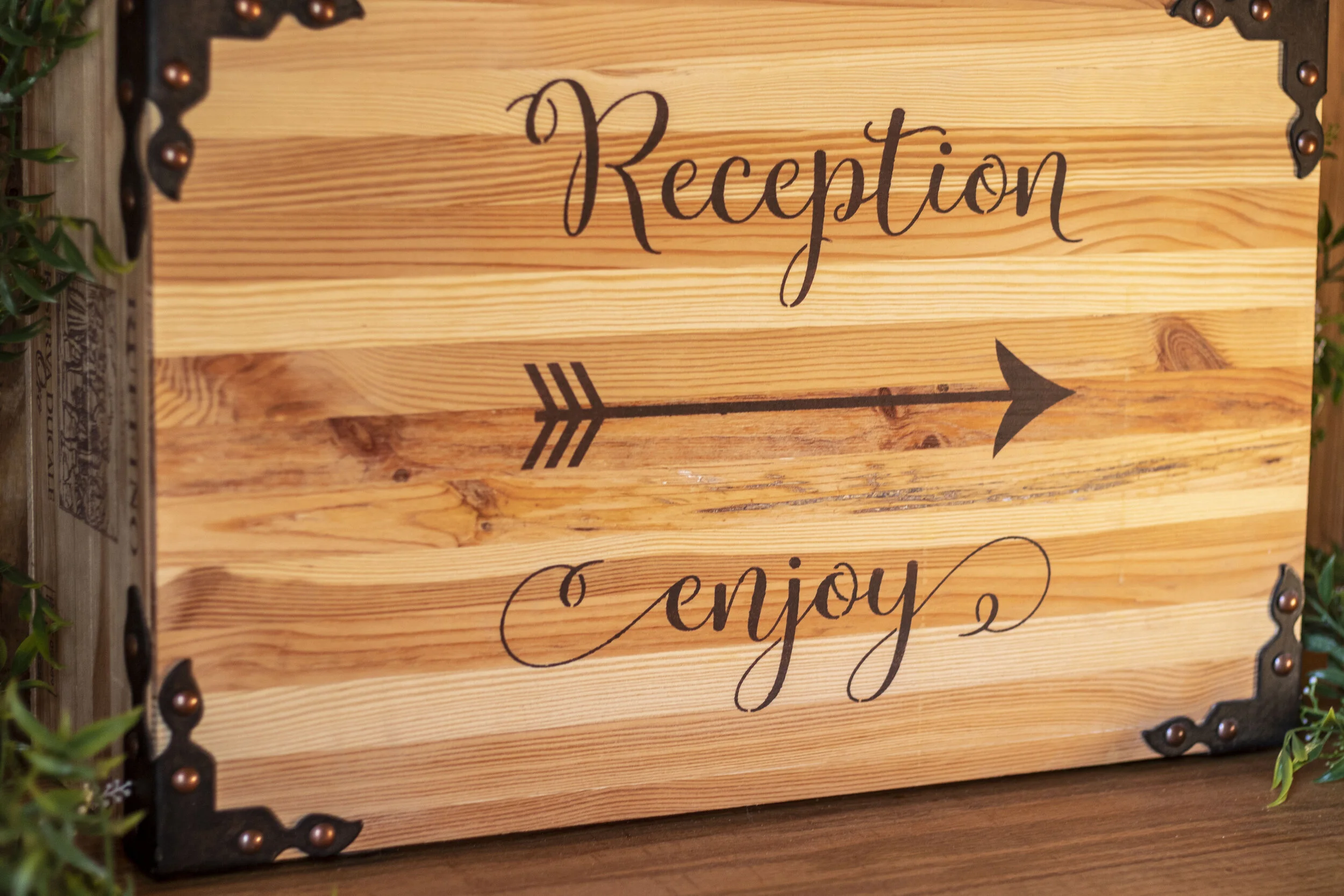 Wooden sign with the words "Reception" and "Enjoy" and a decorative arrow, bordered with metal corner accents.