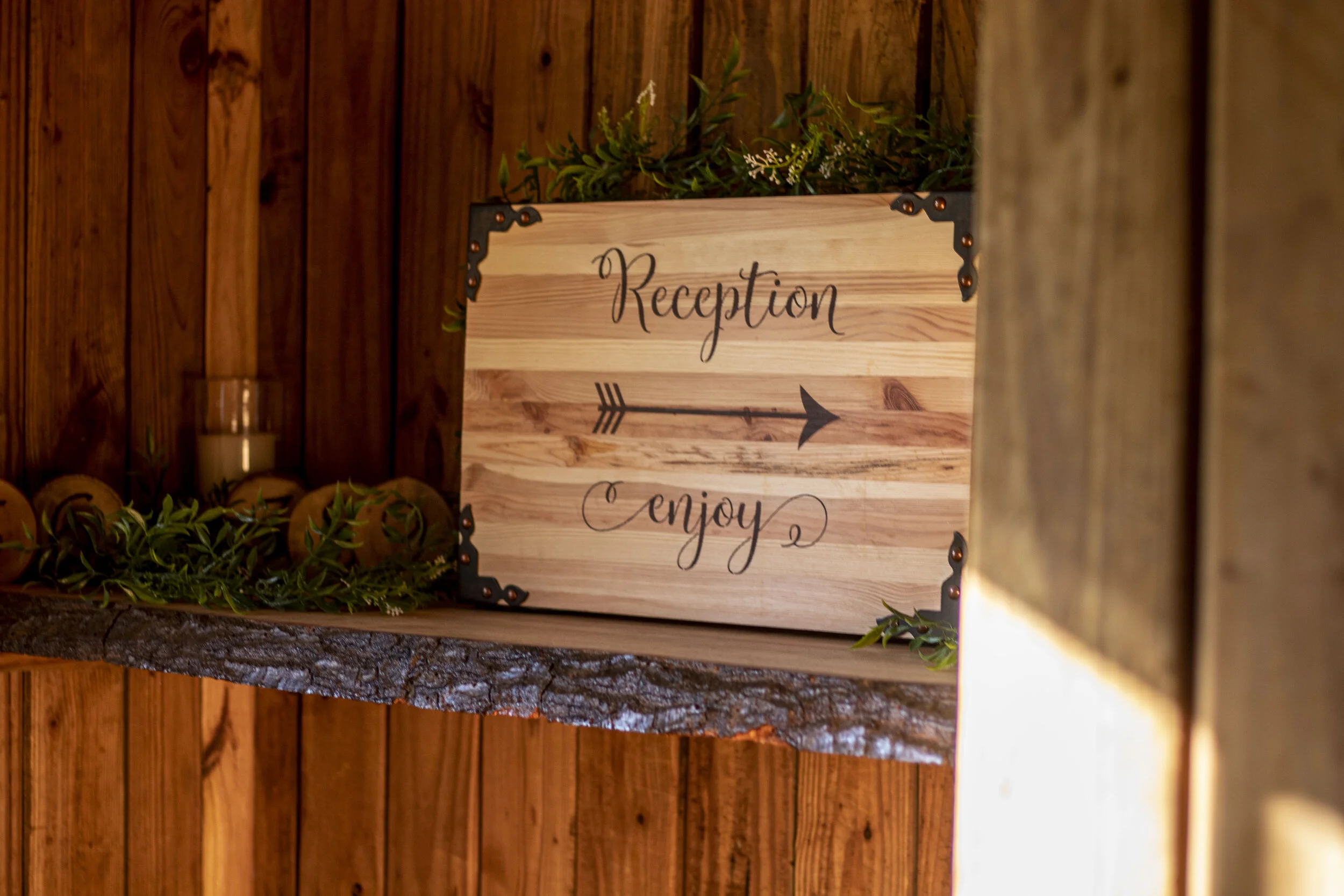 Wooden reception sign with text "Reception Enjoy," decorated with an arrow and surrounded by greenery on a rustic shelf.