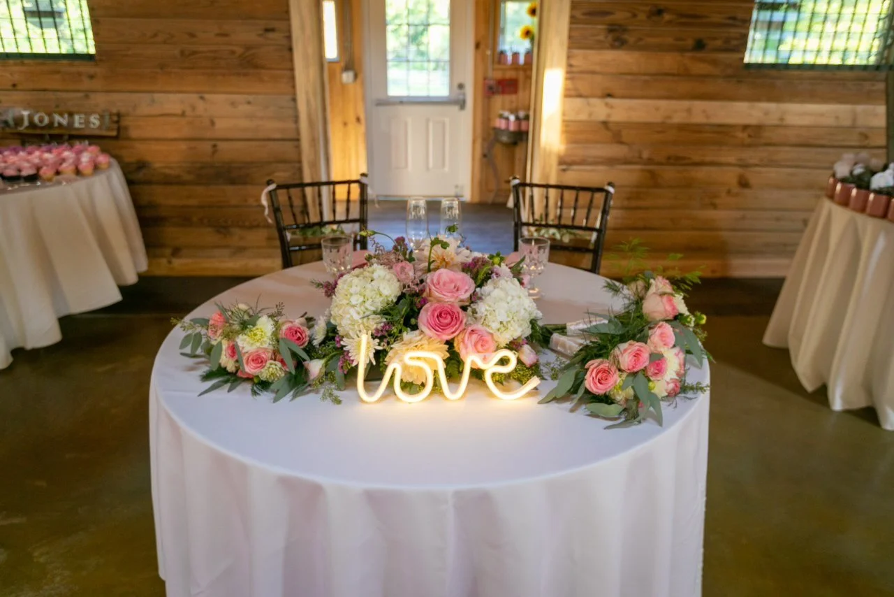 Wedding sweetheart table with floral arrangement, "love" sign, wooden backdrop.