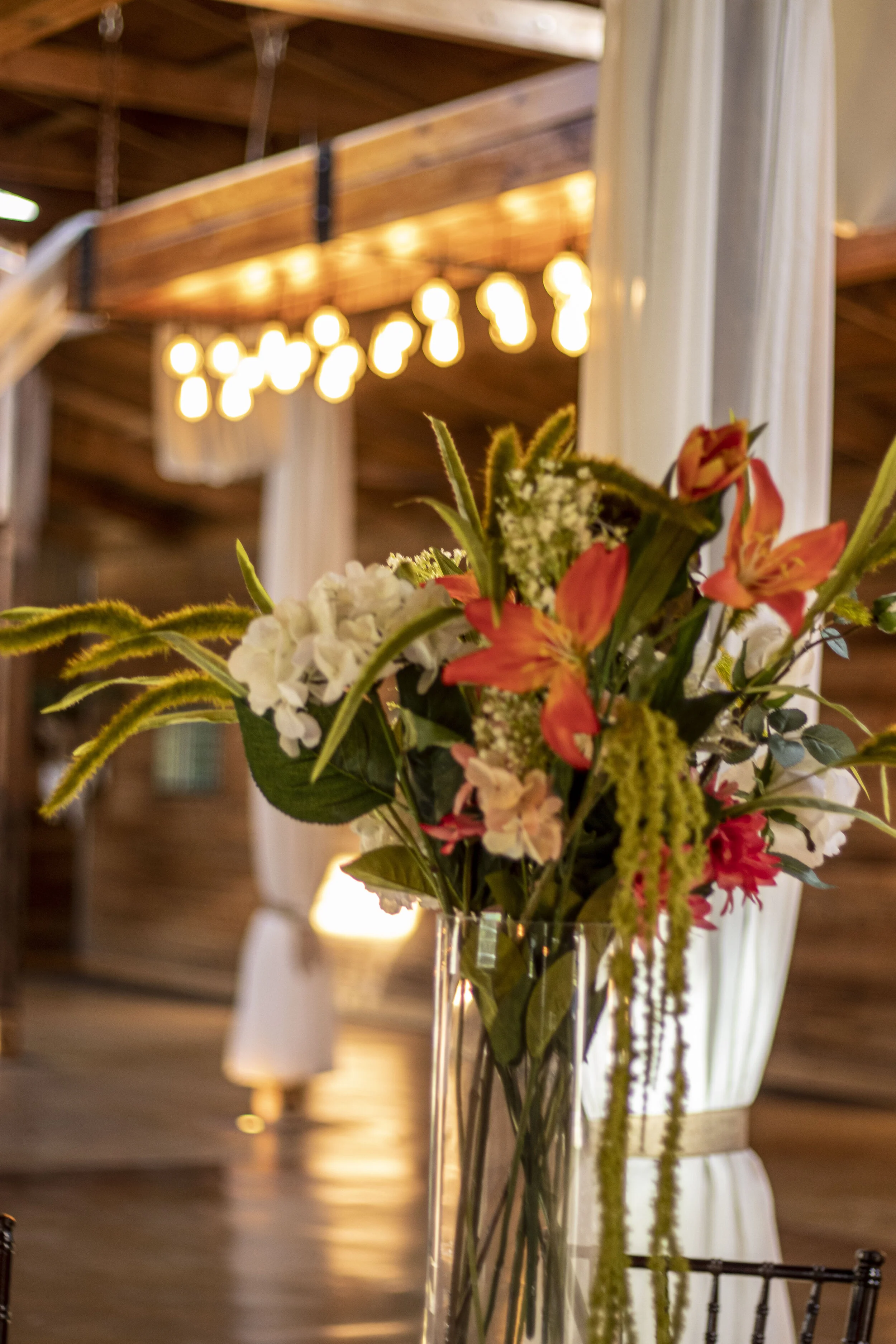 Floral arrangement with orange lilies and white hydrangeas in a glass vase in a rustic setting with draped white curtains and warm lighting.