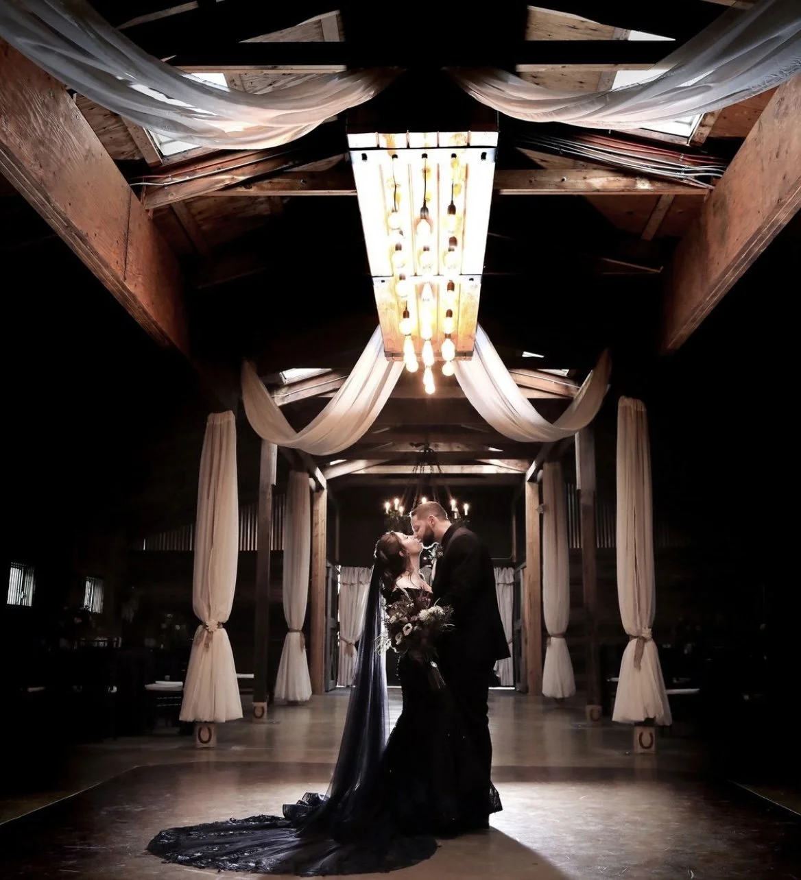 A couple in formal attire stands in a dimly lit venue with draped fabric and chandeliers, sharing an intimate moment. The setting appears elegant and romantic.
