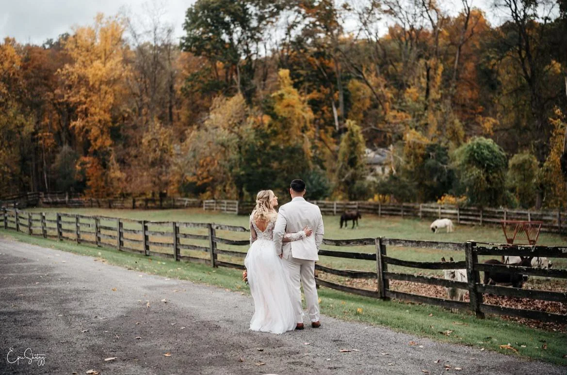 A couple in wedding attire walking arm-in-arm on a path beside a wooden fence with a field and autumn trees in the background.