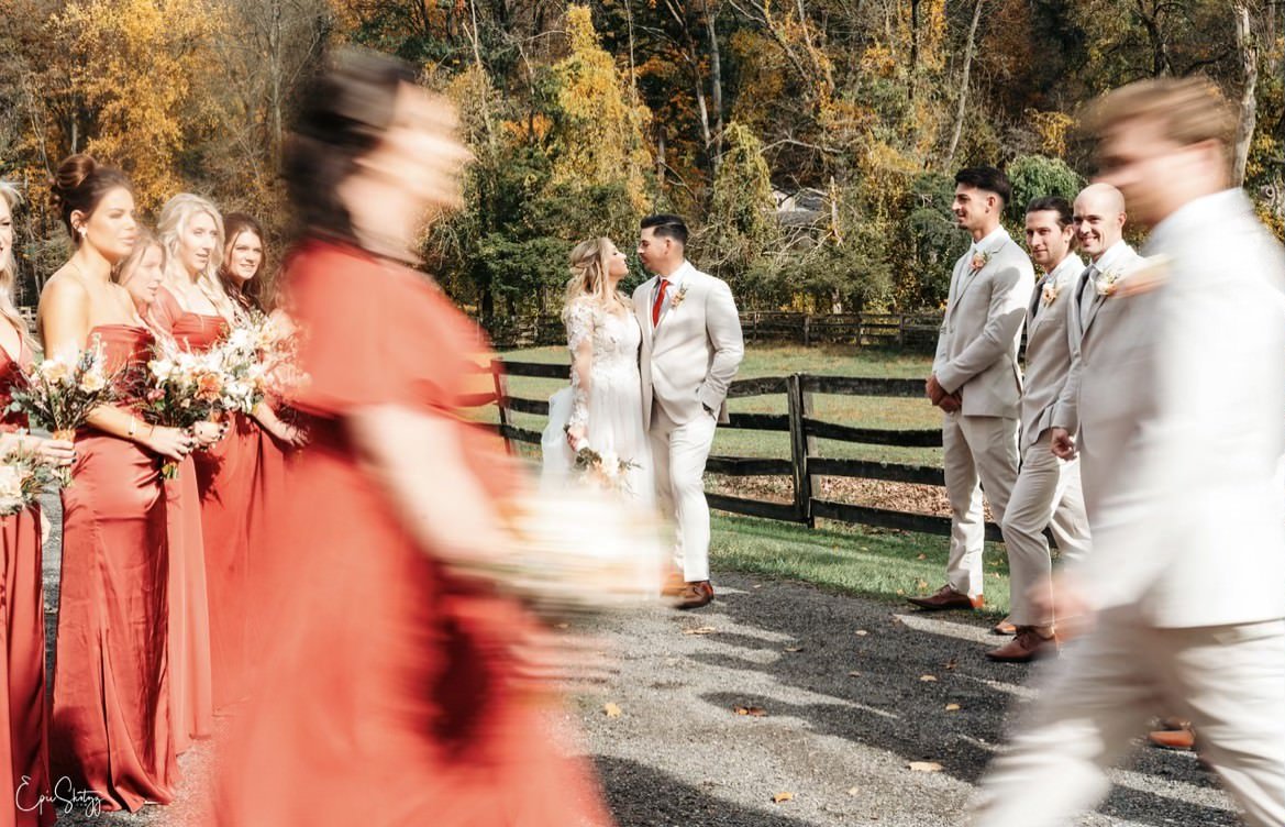 Wedding ceremony with bride and groom, bridesmaids in red dresses, groomsmen in light suits, blurred figures in foreground, outdoor setting with fall foliage.