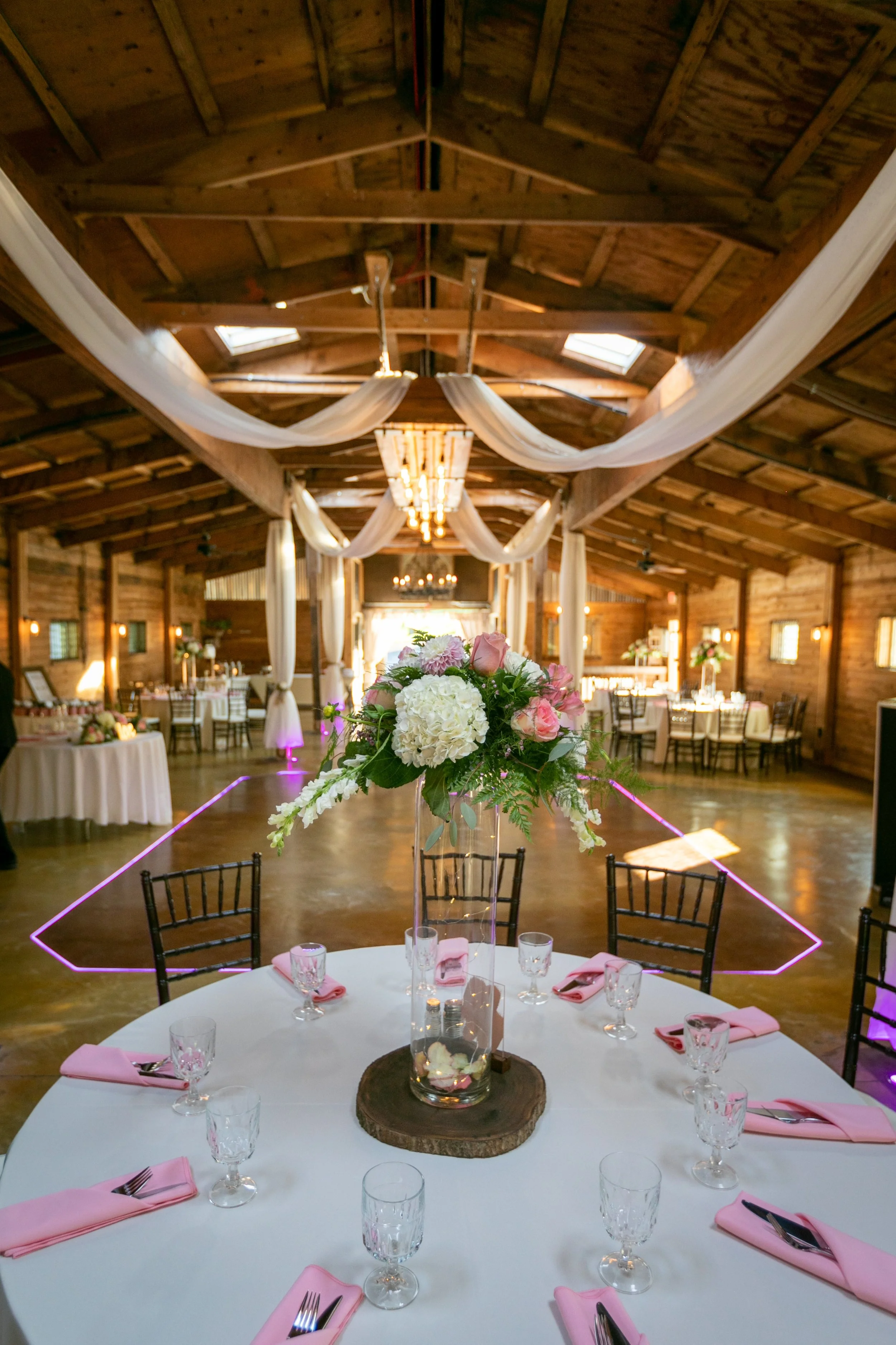 Rustic wedding reception venue with wooden beams, draped white fabric, and a round table set with pink napkins and glassware. A floral centerpiece with roses and hydrangeas is on the table.