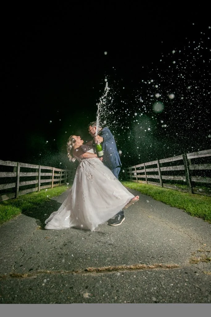 Bride and groom celebrating at night, popping a champagne bottle with splashes in the air, dressed in wedding attire, standing on a path with fences.