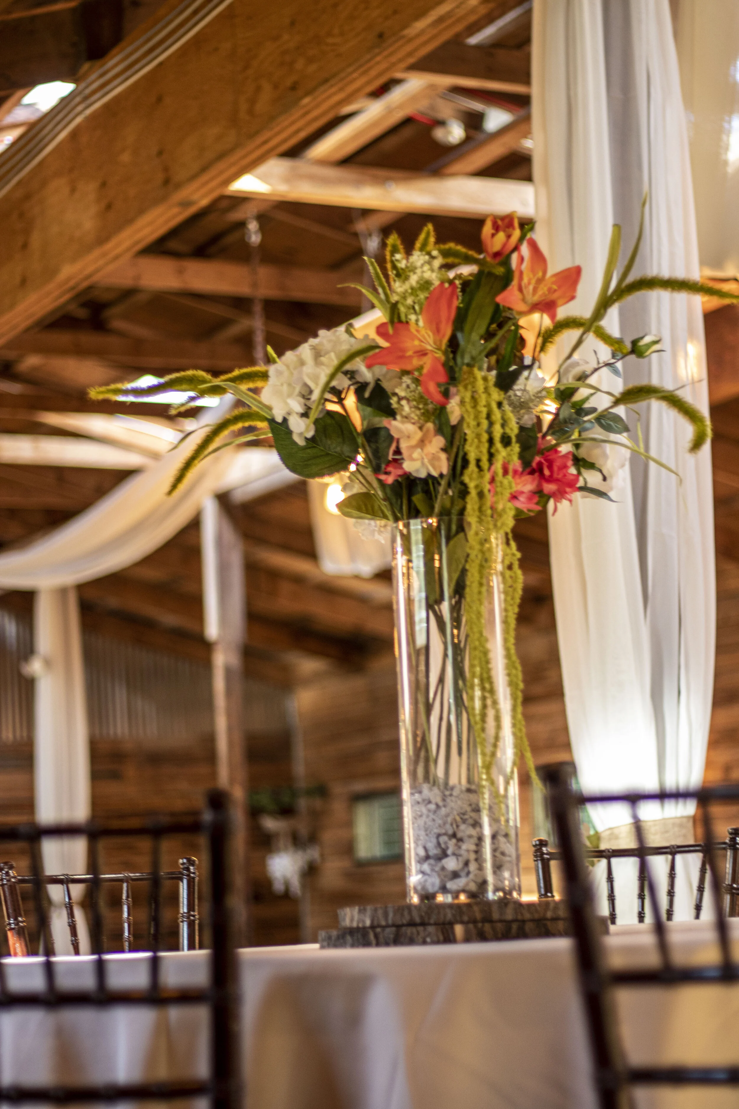 Floral centerpiece with lilies and white flowers in a tall vase at a rustic venue.