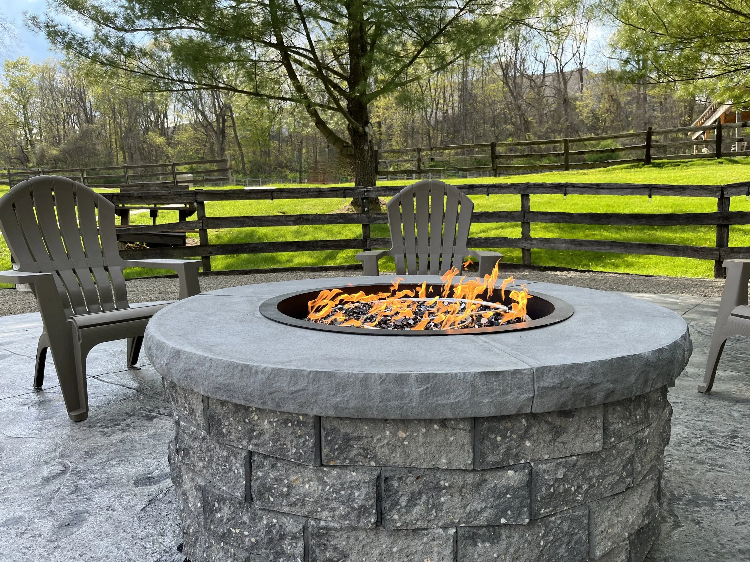 Stone fire pit with orange flames surrounded by three gray Adirondack chairs, set on a patio with a wooden fence and green lawn in the background.