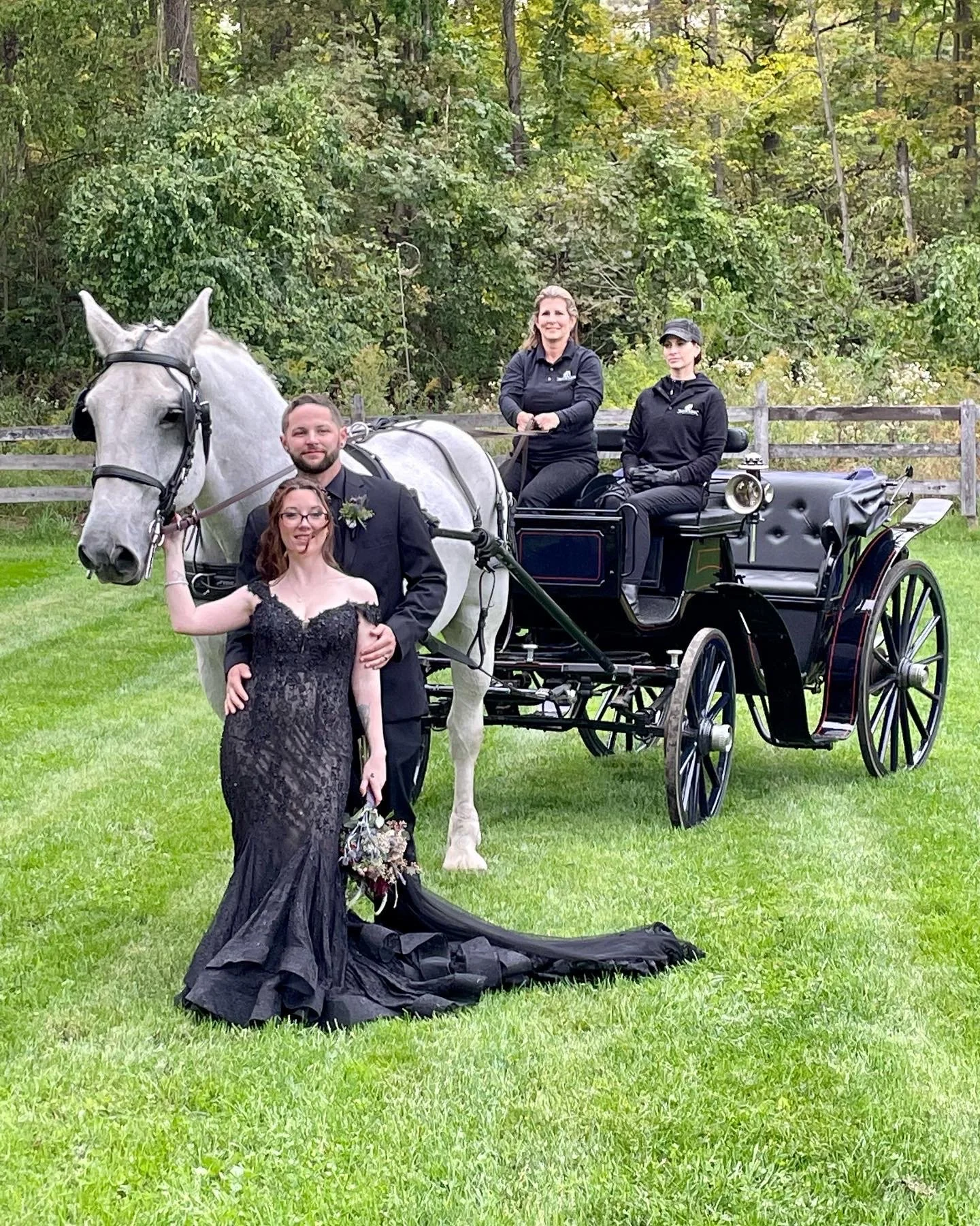 A couple in formal attire poses with a white horse and carriage. The woman wears a black lace gown, and the man is in a black suit. Two people are seated on the carriage. The setting is a lush, green outdoor area.