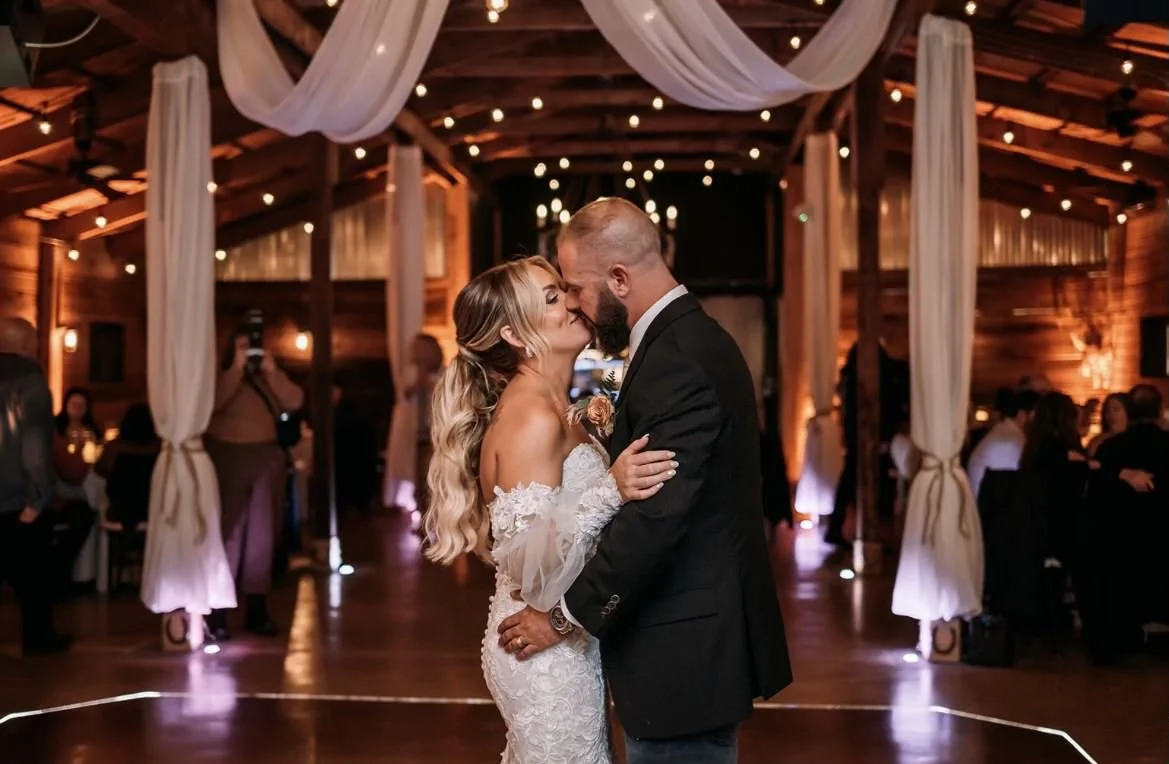 Bride and groom embracing during their first dance at a rustic wedding venue with wooden beams and string lights.