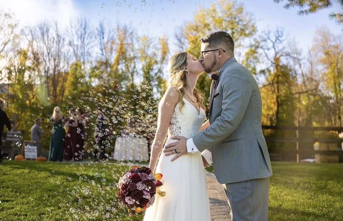 Bride and groom kissing outdoors surrounded by bubbles and autumn trees.