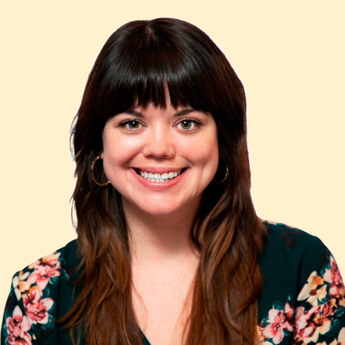 A woman with long brown hair, wearing hoop earrings, a gold nose ring, and a floral top, smiling at the camera against a light beige background.