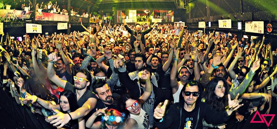 Crowd of people at a concert or rave event, dancing and enjoying music, with colorful lighting and some wearing sunglasses or accessories.
