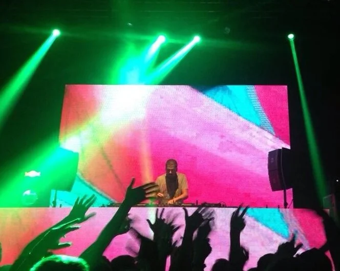 A DJ performing on stage with a large colorful LED screen behind them, bright green stage lights, and an enthusiastic crowd with raised hands.