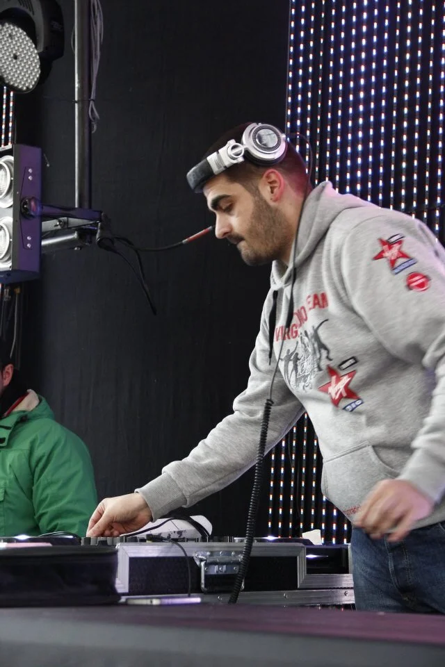 A man with a beard wearing a gray hoodie and headphones is DJing at a music event. He is adjusting equipment on a table, with a person in a green jacket partially visible on his left. The background features a black wall and a vertical LED light disp