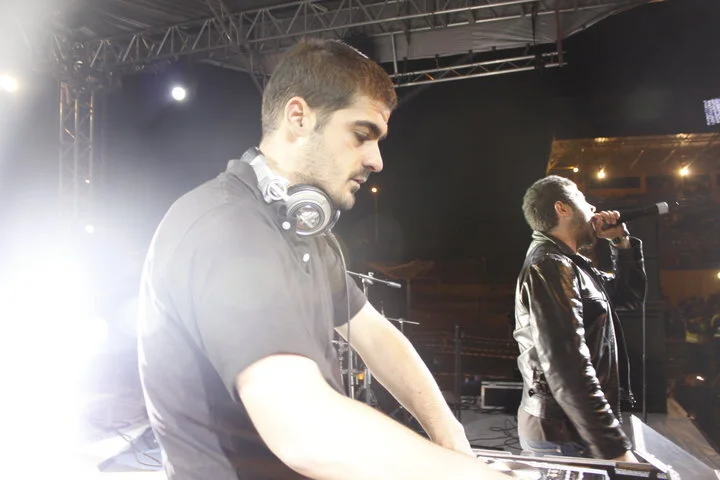 Two male performers on stage, one with headphones around his neck, operating DJ equipment, the other singing into a microphone, with stage lighting and equipment in the background.