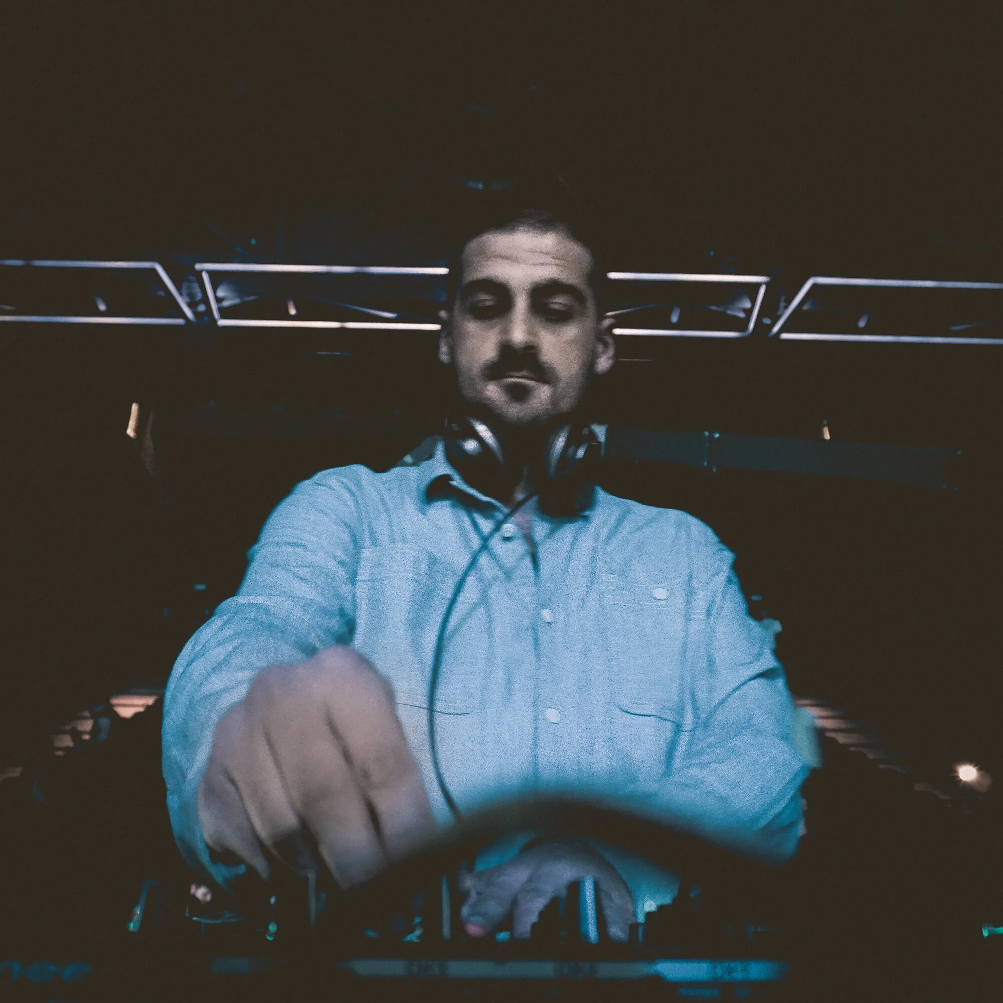 A male DJ with headphones around his neck is DJing at a nightclub or event with stage lighting in the background.