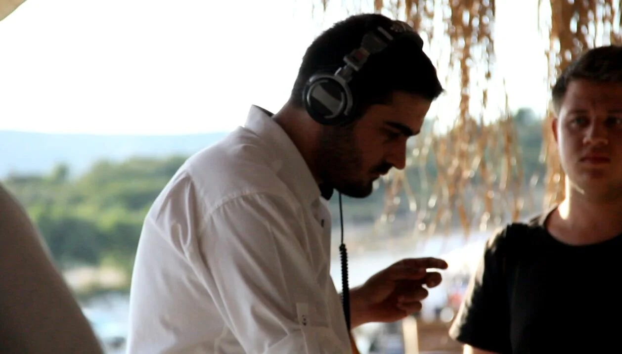 Man wearing headphones and a white shirt, leaning forward with a focused expression, with a young man standing nearby in an outdoor setting with trees and water in the background.
