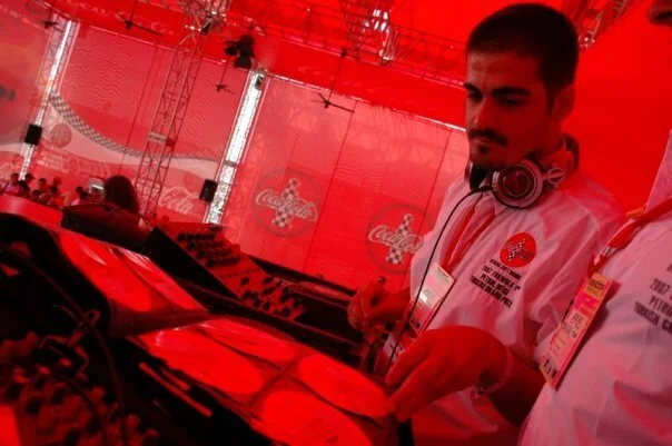 Two DJs wearing white shirts with a Coca-Cola logo, working with DJ equipment under a red canopy at an event.