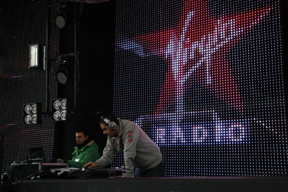 Two DJs wearing headphones, working at a DJ booth with a large LED screen in the background displaying a stylized logo.