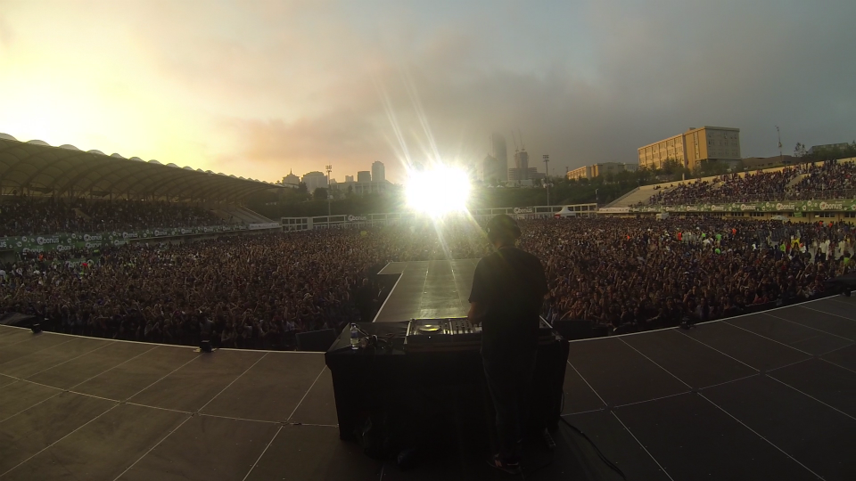 A DJ stands behind turntables on stage at a concert, facing a large crowd in an outdoor stadium, with city buildings and a sunset sky in the background.