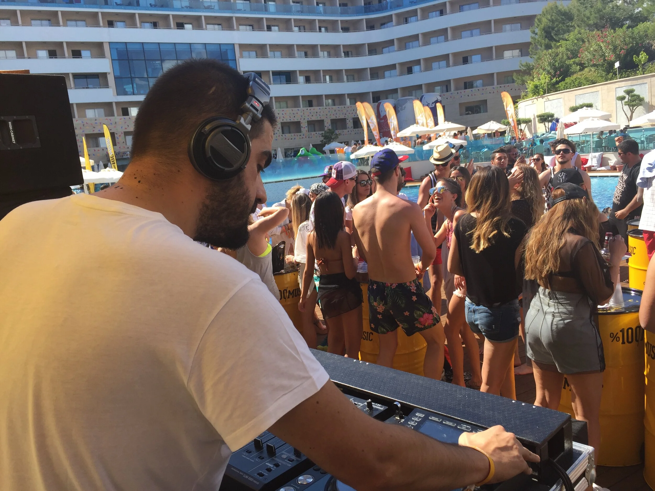 DJ wearing headphones at poolside party with people dancing and socializing, pool and hotel building in background.