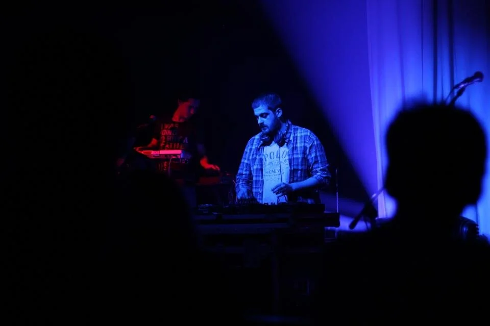 A DJ is performing with blue lighting on stage, with a person in the foreground watching. The DJ is wearing a plaid shirt and headphones.