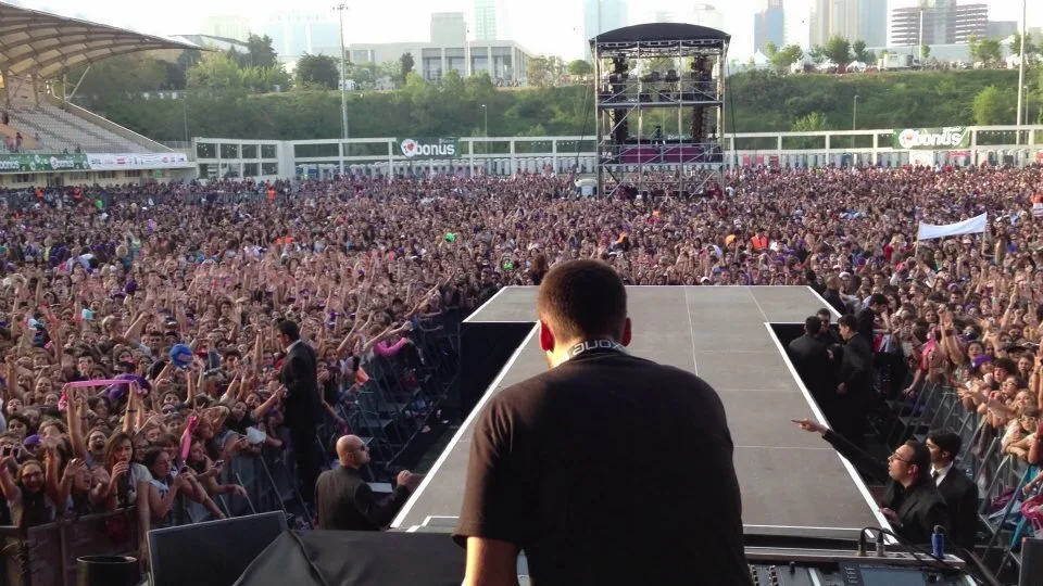 A large outdoor concert with a massive crowd facing a stage, seen from behind a performer or DJ at the front with DJ equipment.