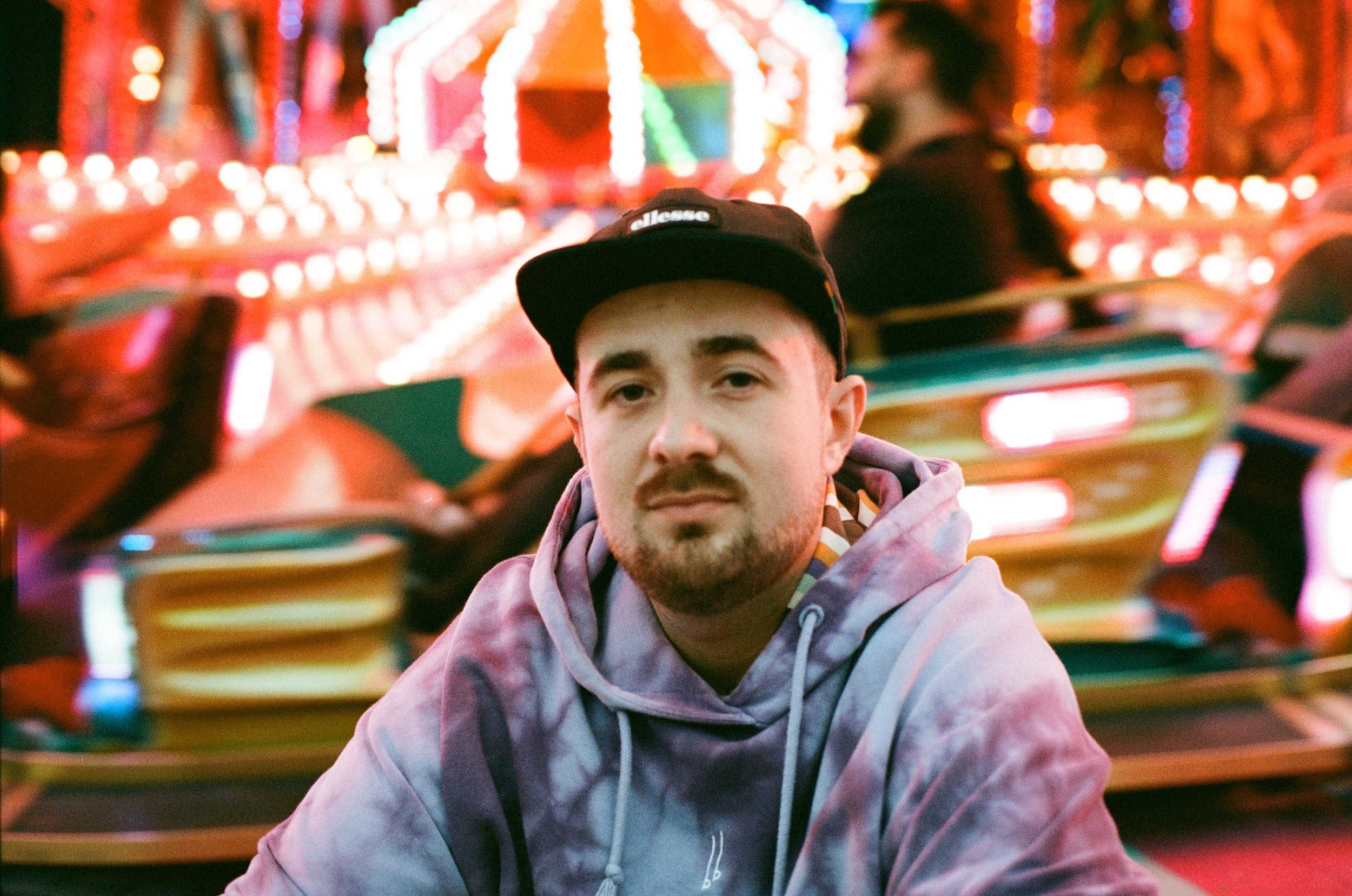 Ein Mann sitzt vor einem Karussell bei einem Jahrmarkt, er trägt eine schwarze Kappe und einen Hoodie, im Hintergrund bunte, leuchtende Karusselllichter.