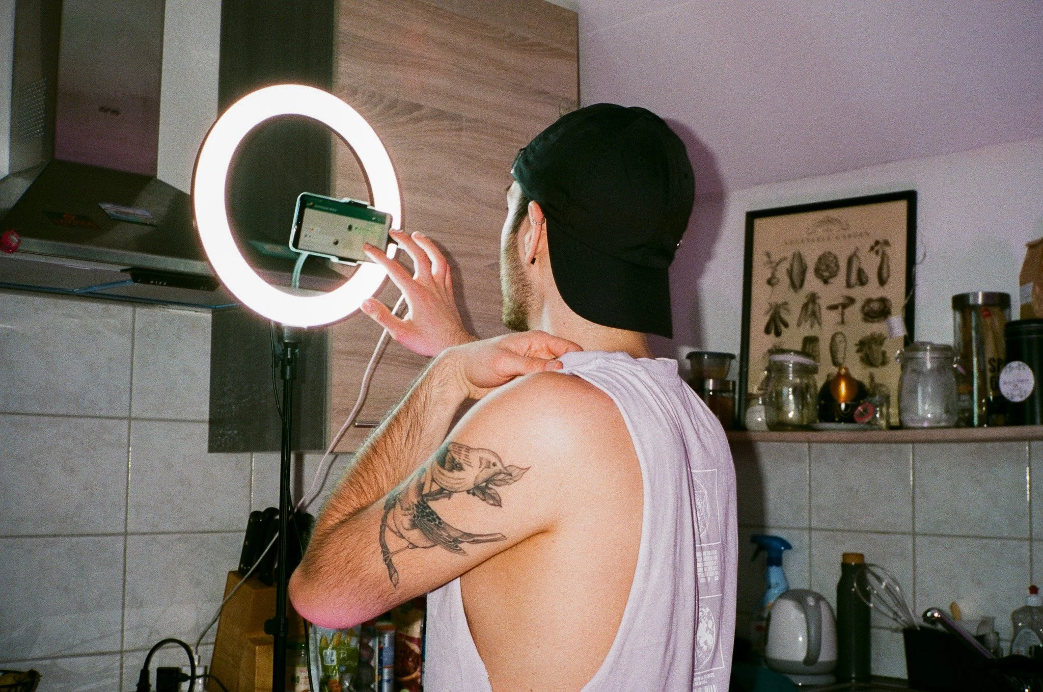 Jemand macht ein Selfie vor einem beleuchteten Spiegel in einer Küchenumgebung, mit Tattoos am Arm, trägt eine schwarze Kappe und ein ärmelloses T-Shirt.