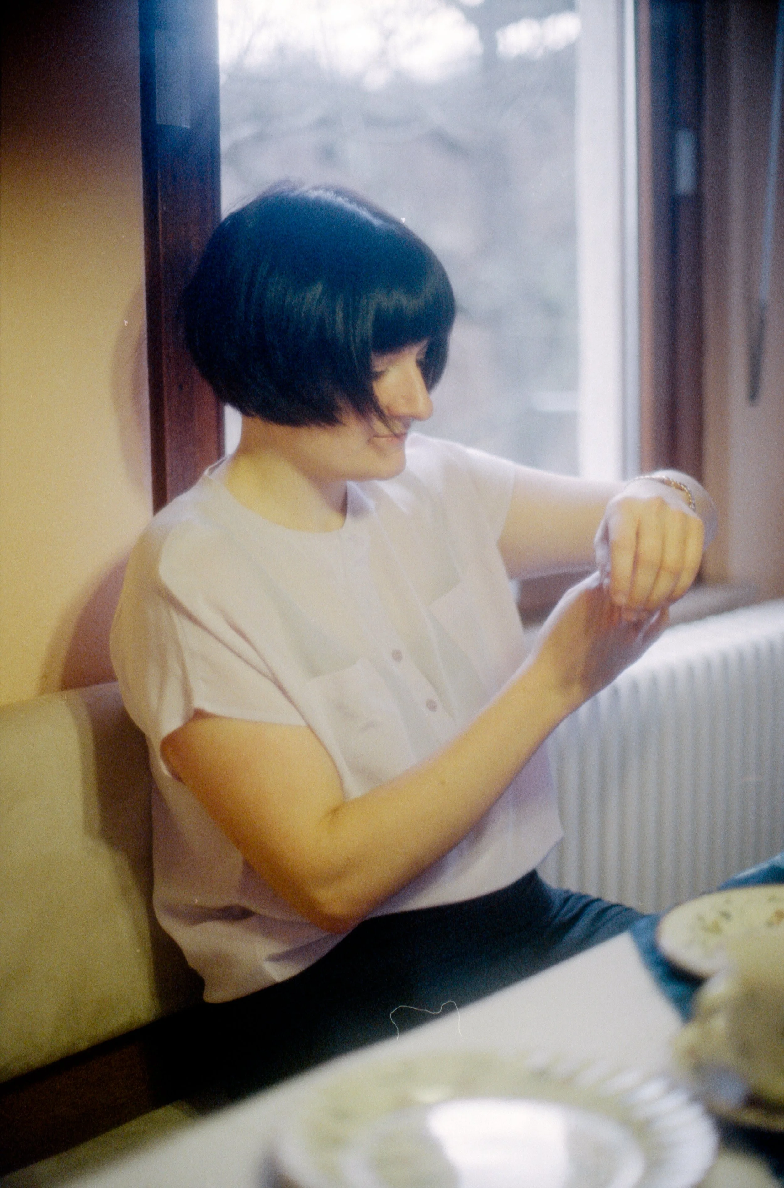 Eine Frau mit schwarzen Haarn und weißem Hemd sitzt am Tisch vor einem Fenster, schaut auf ihre Armbanduhr und lächelt, während sie die Uhr betrachtet.