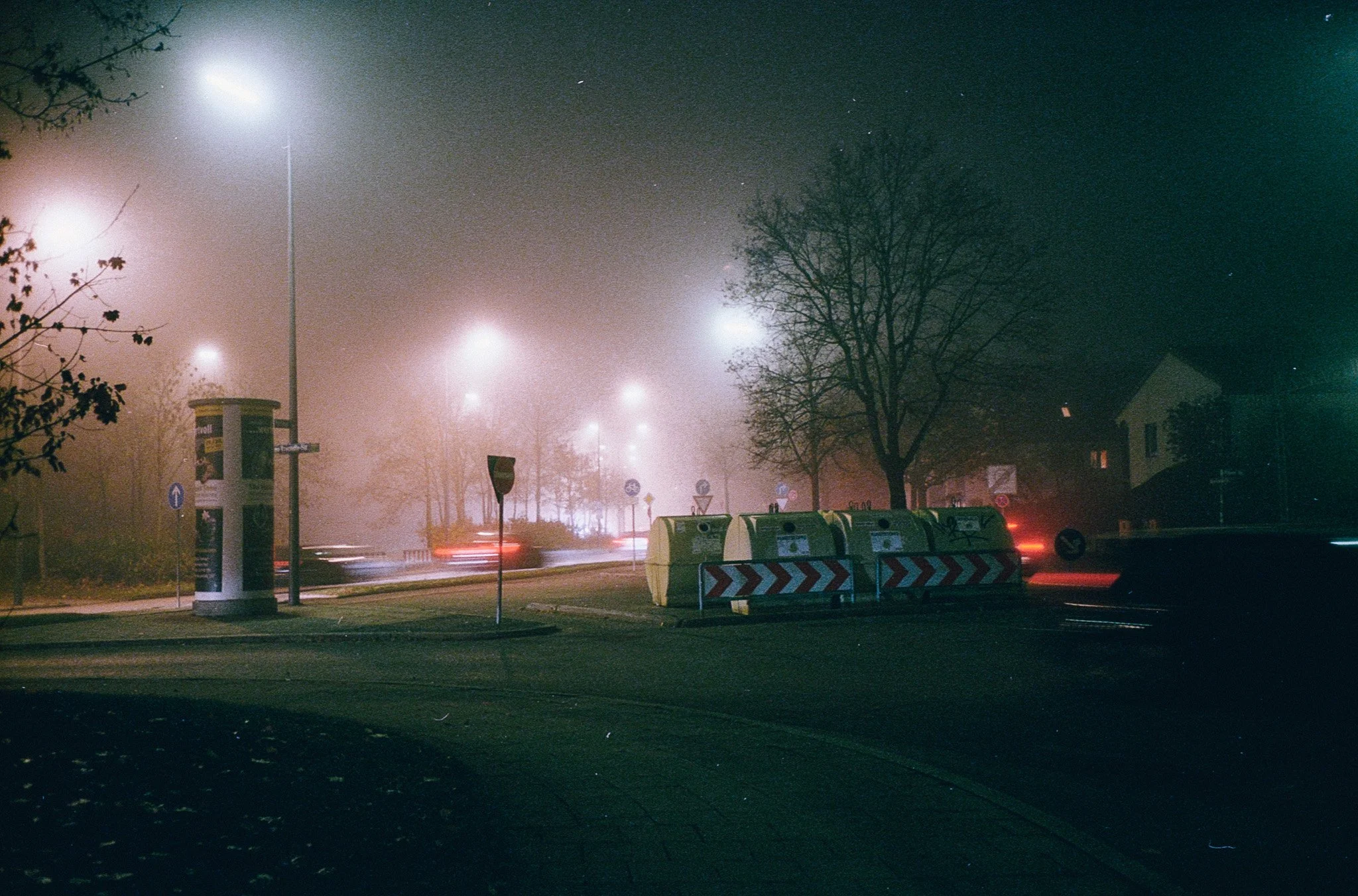 Nebelverhüllte Straßenlaternen leuchten in einer nächtlichen, nebligen Straße, mit leerem Straßenrand, Fahrradständern, einem Telefonkiosk und einem Baum, im Hintergrund sind Häuser sichtbar.