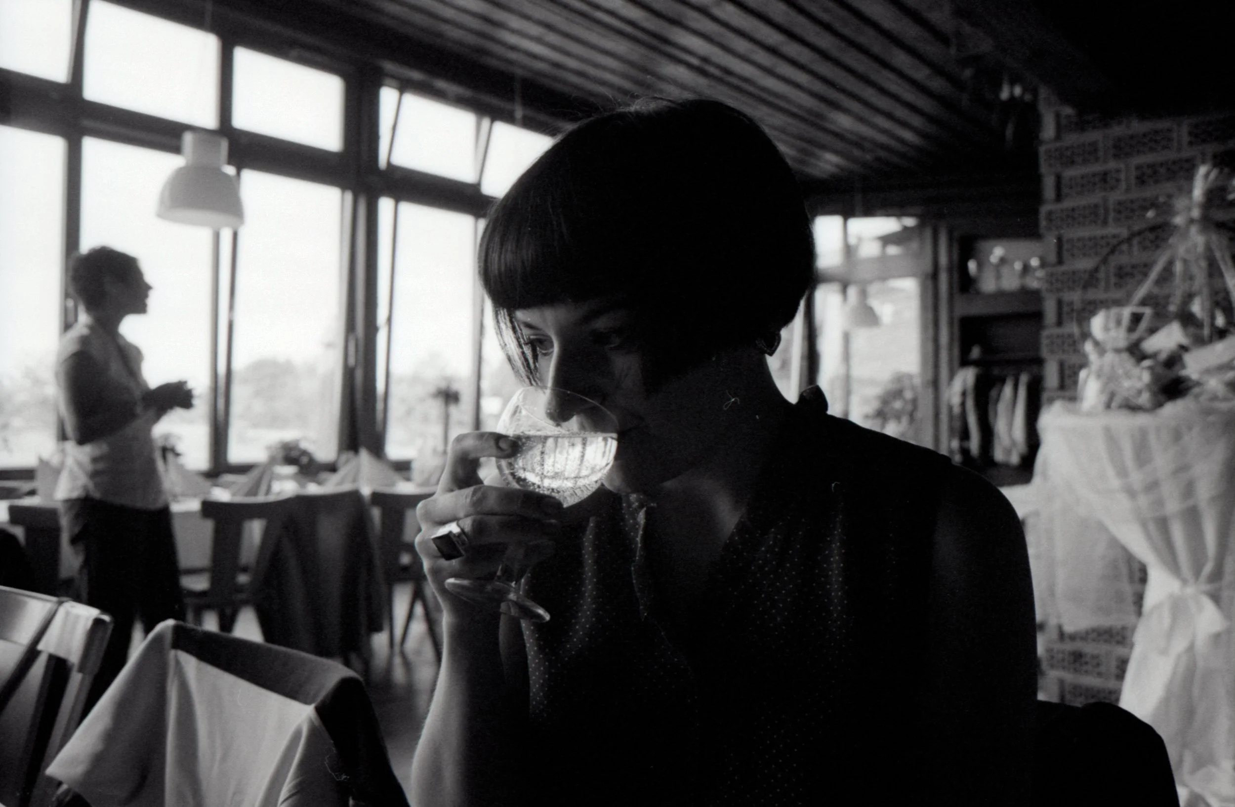 Schwarz-weiß Foto einer Frau mit kurzem Haar, die ein Weinglas hält und daraus trinkt, in einem Restaurant mit großen Fenstern im Hintergrund.