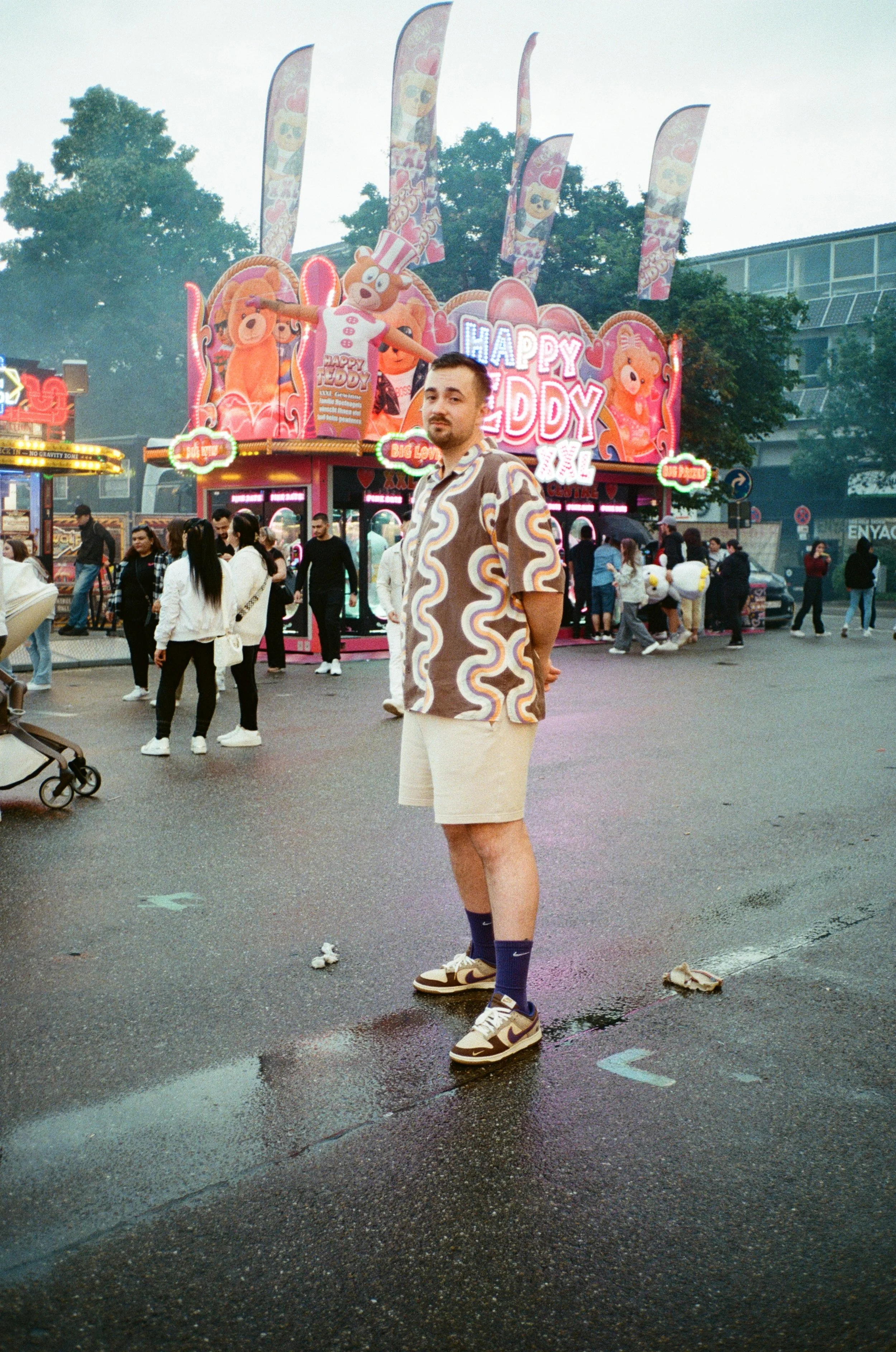 Junger Mann in buntem Hemd und Shorts steht vor einem Jahrmarkt mit Kinderfahrgeschäften. Im Hintergrund ist ein Karussell mit der Aufschrift 'Happy Teddy XXL' und bunten Banner. Es ist bewölkt und die Straße ist nass.