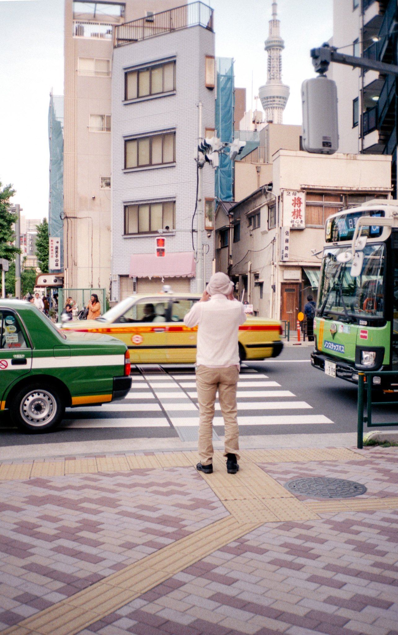 Ein älterer Mann steht an der Straßenkreuzung und fotografiert vorbeifahrende Autos, während im Hintergrund Gebäude und der Fernsehturm sichtbar sind.