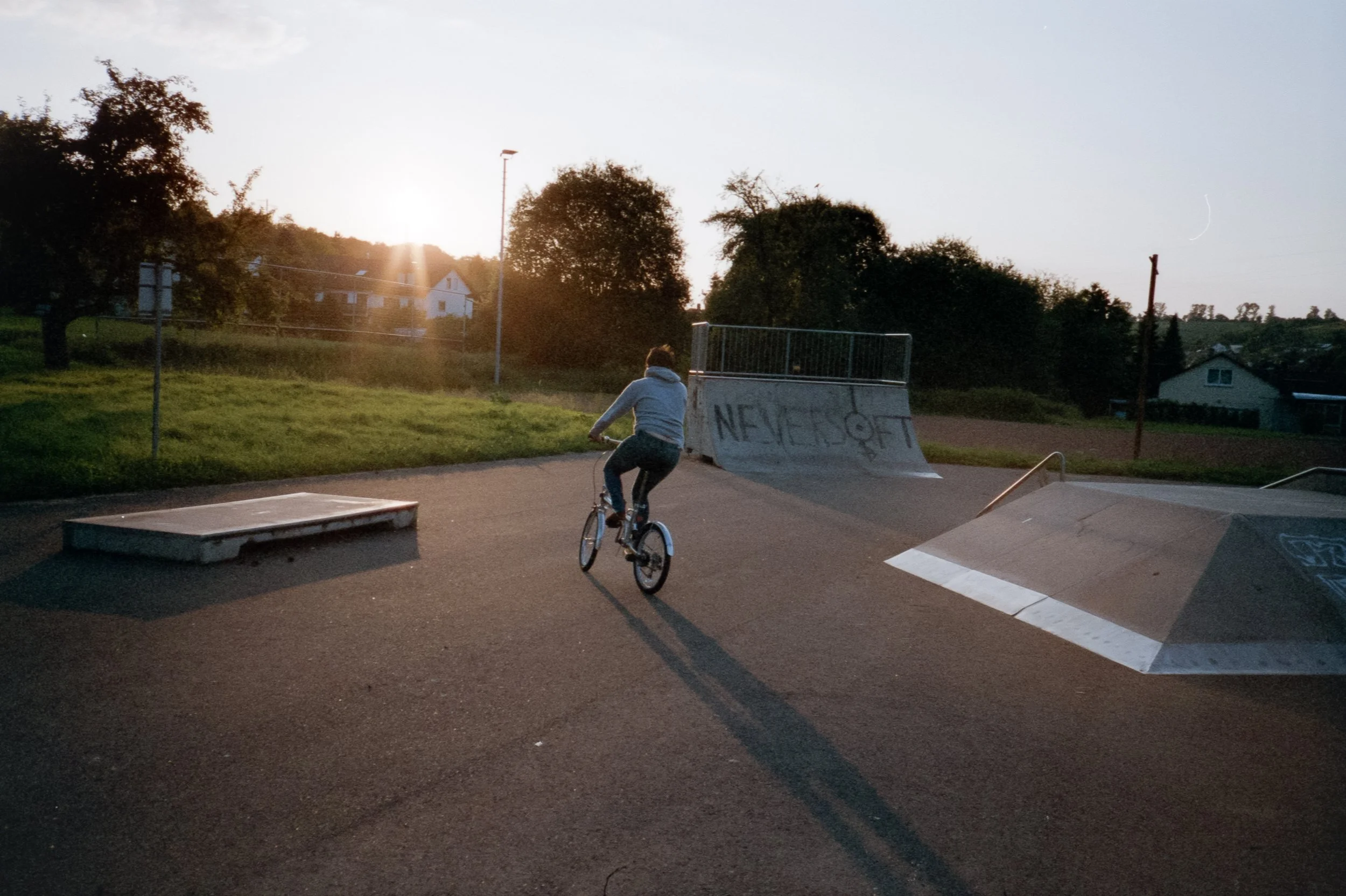 Junge fährt Fahrrad auf Skatepark bei Sonnenuntergang, mit graffiti-beschriebenem Sprunggerät im Hintergrund.