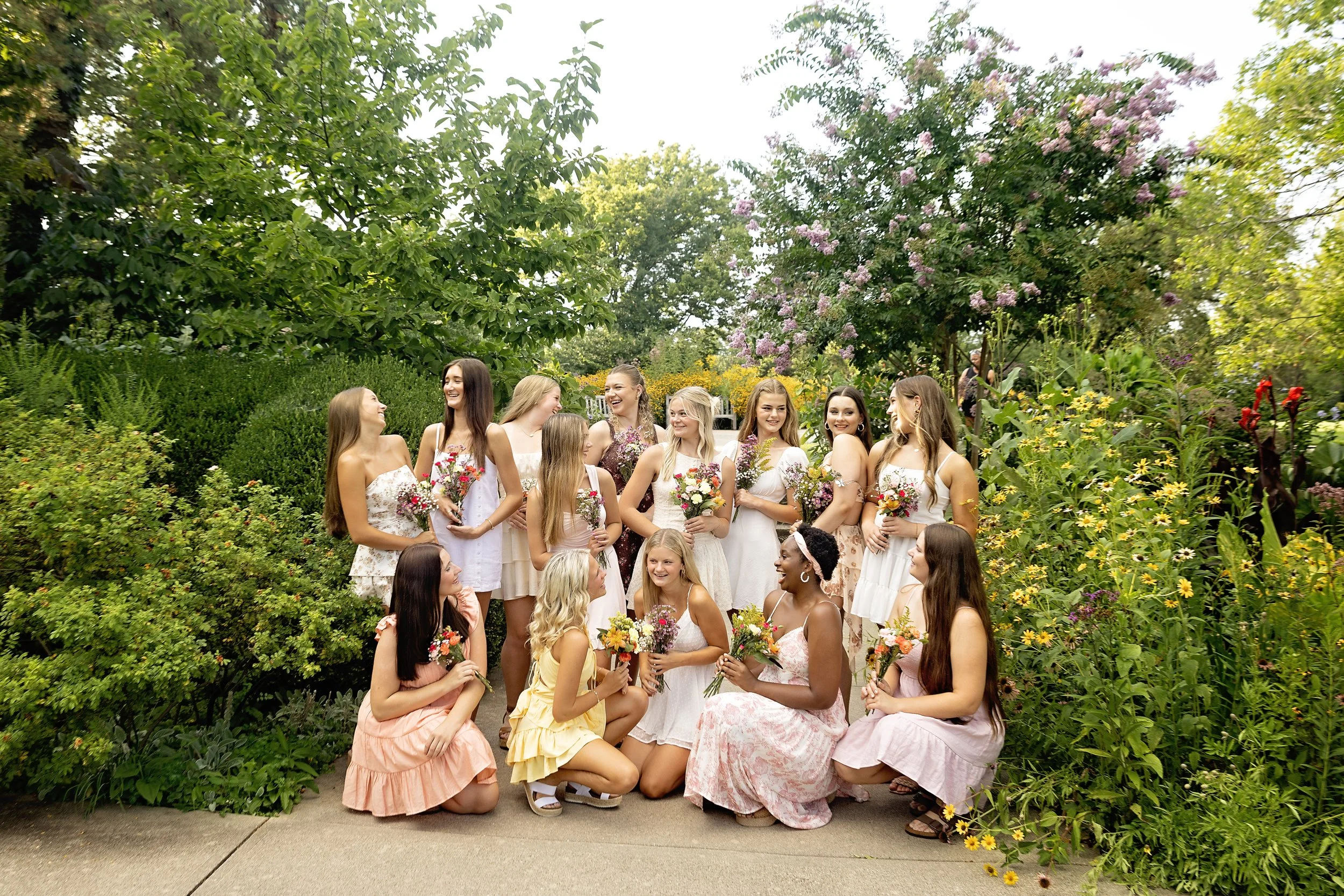 A group of young women and girls dressed in summer dresses, holding colorful bouquets, gathered outdoors in a lush garden with greenery and blooming flowers, smiling and engaging with each other.