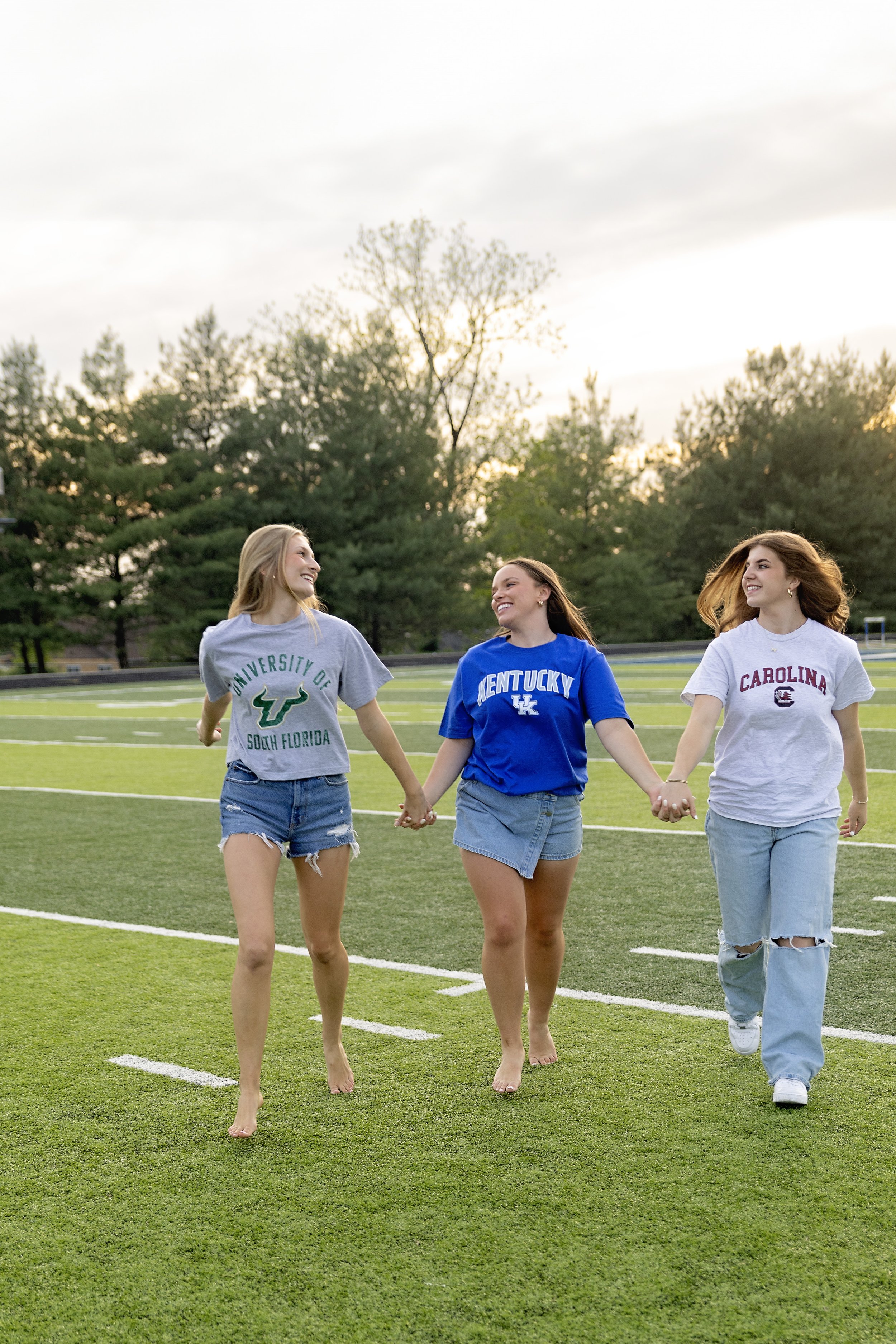 Three young women holding hands while walking barefoot on a football field at sunset, wearing university T-shirts from South Florida, Kentucky, and South Carolina.