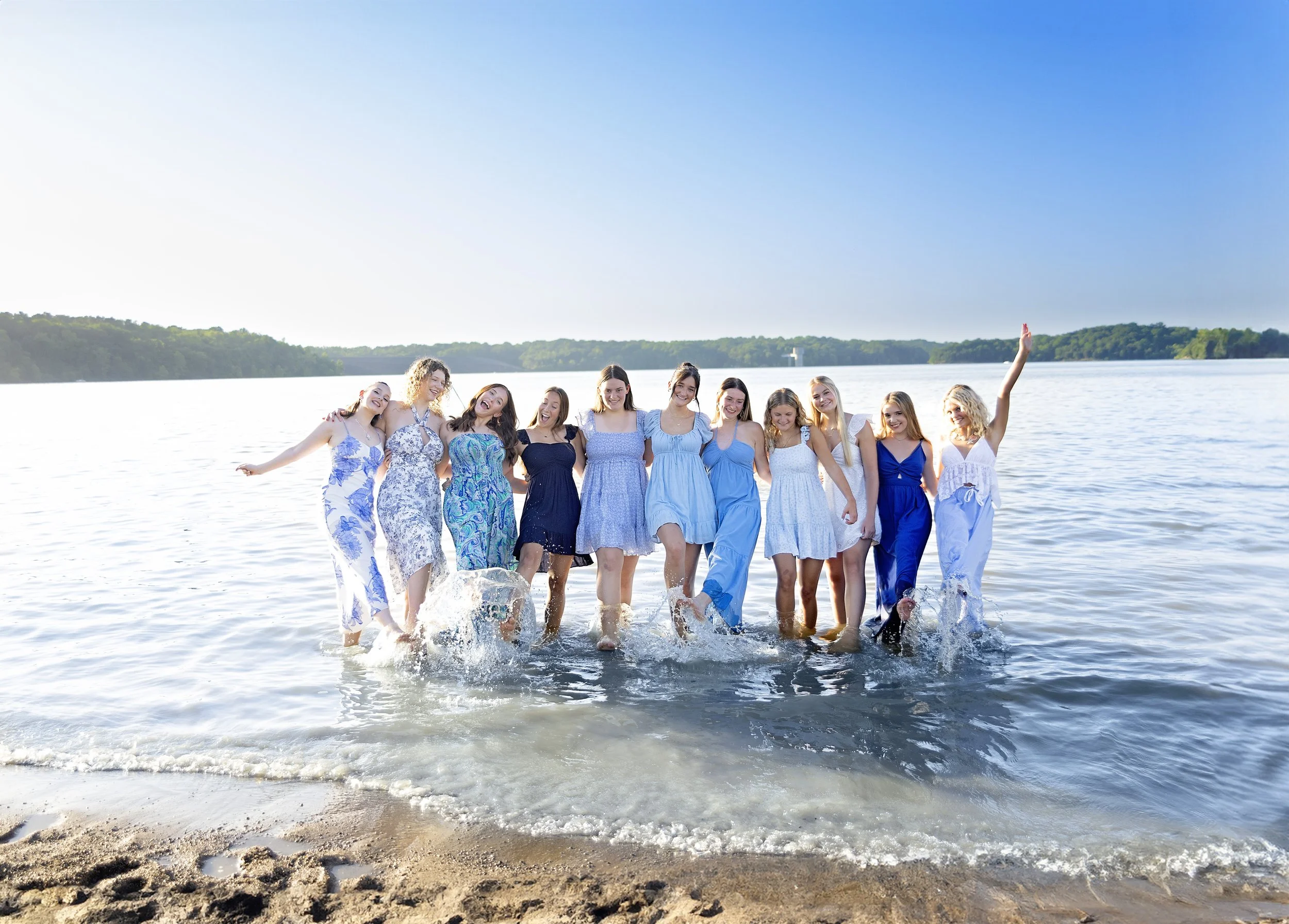 Group of high school seniors in summer dresses standing in shallow water at the beach, smiling and enjoying a sunny day with a clear blue sky