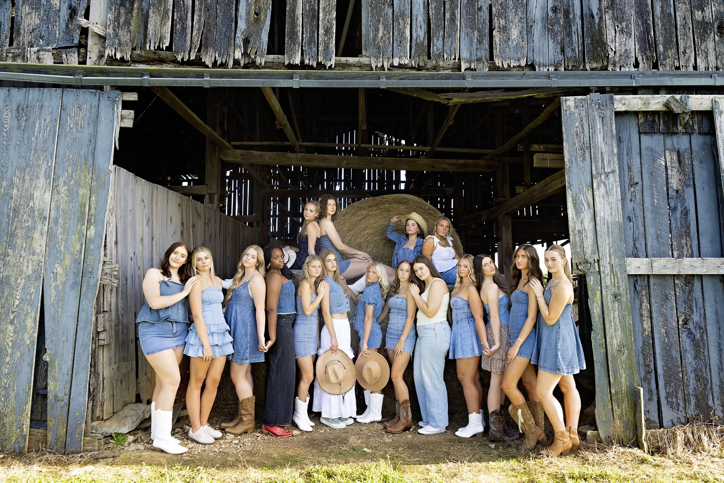 A large group of women are posing inside a rustic barn, with some sitting on a hay bale and others standing. They are wearing denim outfits and cowboy boots, with a couple holding hats. The barn has weathered wood walls and large partially open doors, with sunlight illuminating the scene.