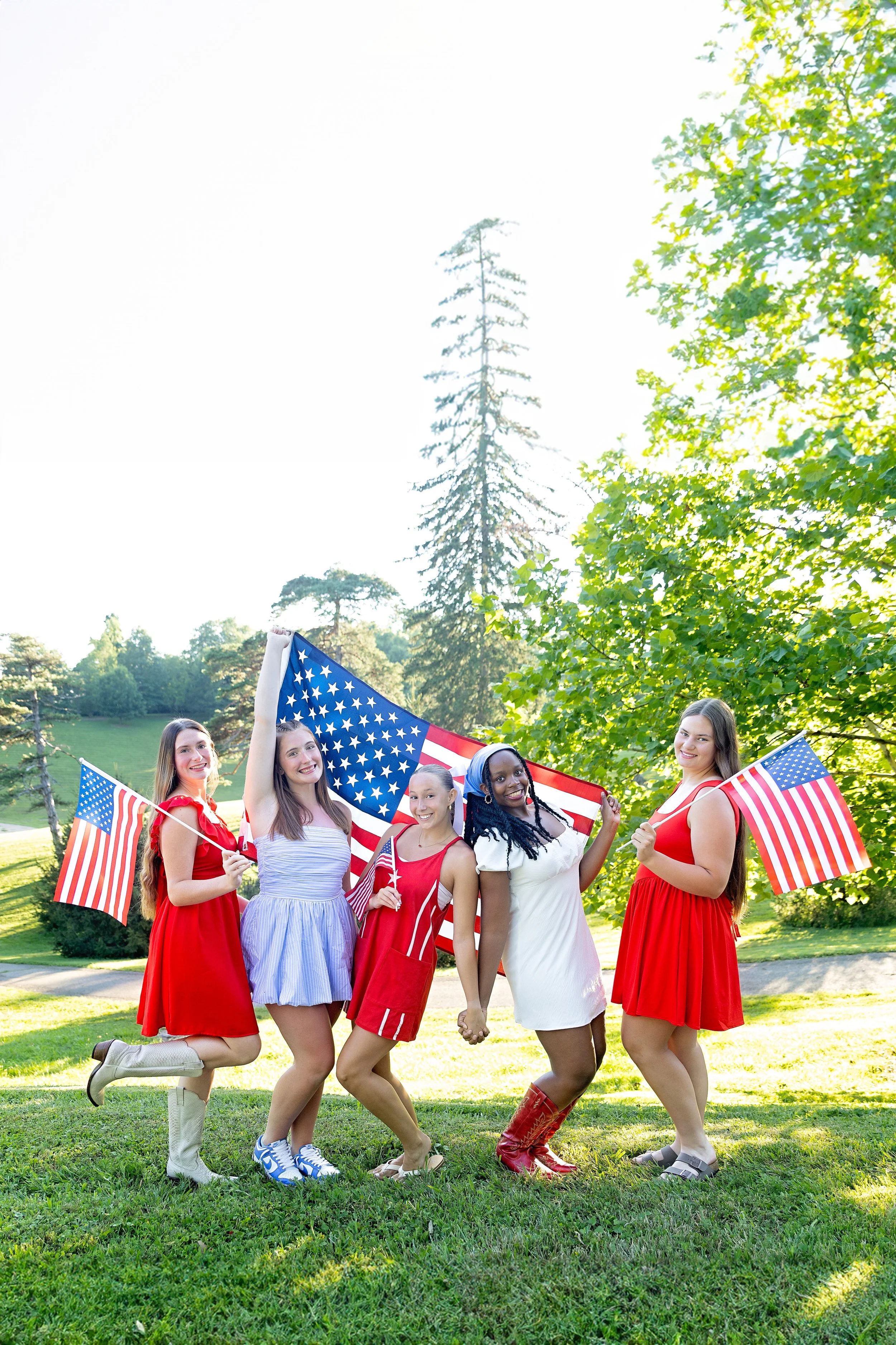 Five women in red, white, and blue clothing celebrating National Independence Day outdoors with American flags, standing on grass with trees and a bright sky in the background.