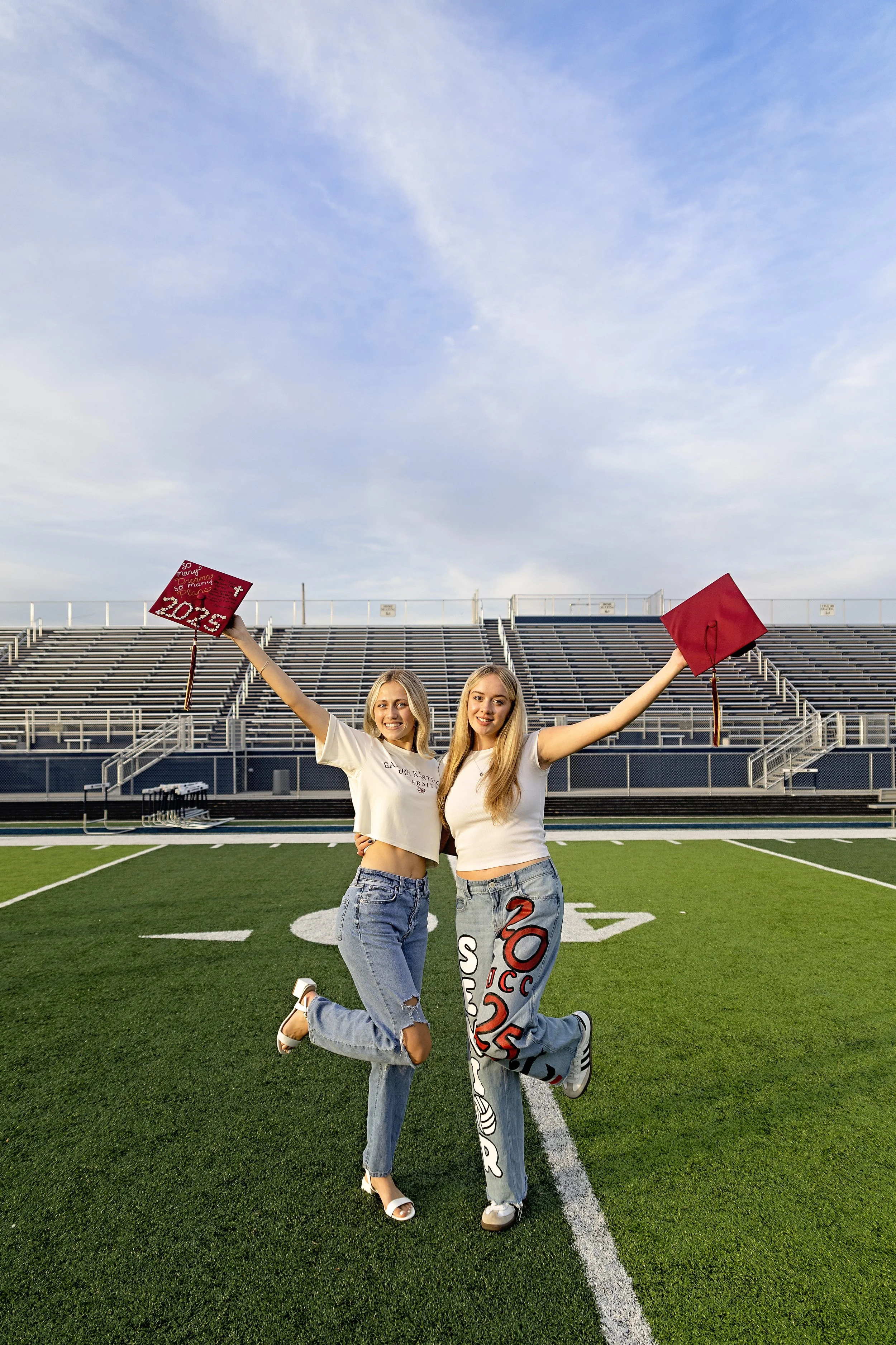 Two young women celebrate graduation on a football field, holding up their graduation caps with outstretched arms.