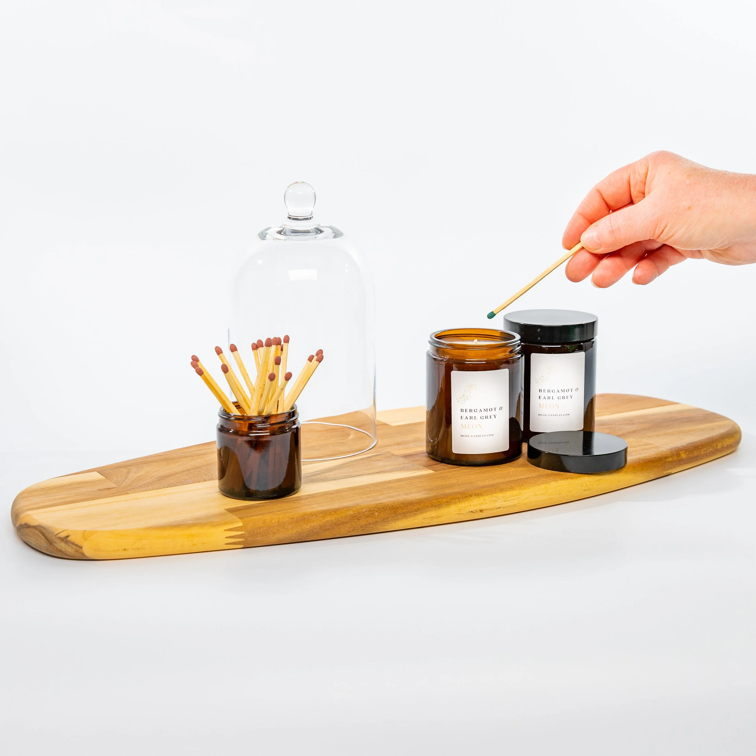 A hand using a matchstick to light a candle in an amber jar on a wooden tray, with another jar of candles, a glass cloche, and a small jar of matches also on the tray.