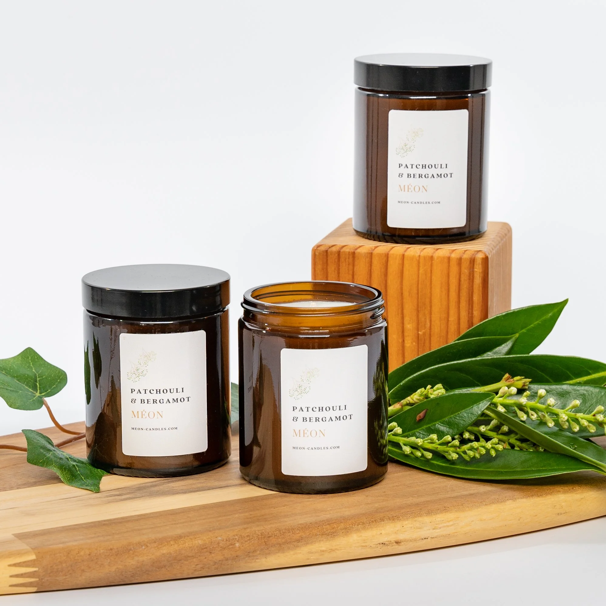 Three amber glass candles with black lids, labeled 'Patchouli & Bergamot Mêon', arranged on a wooden board and a small wooden block, decorated with green leaves and budding white flowers.