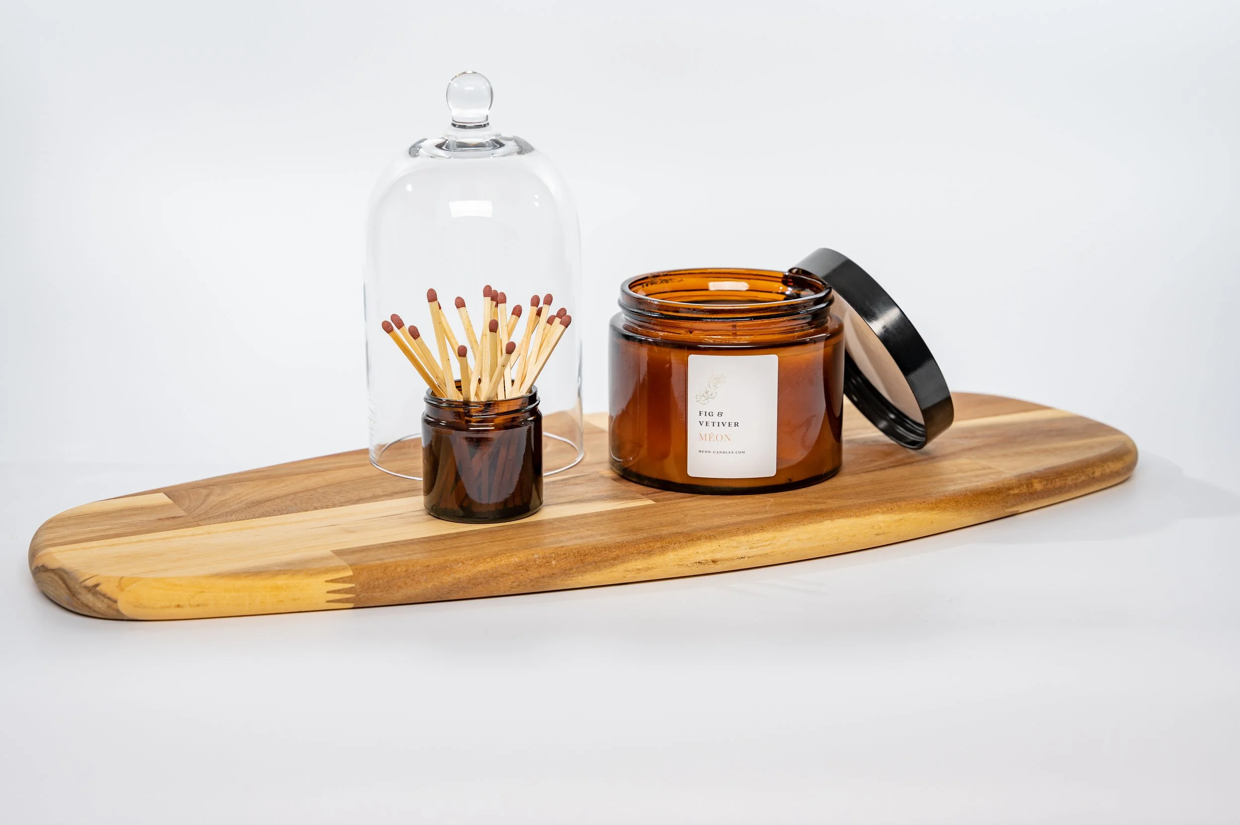 Wooden tray holding a glass cover, a small jar of matches, and a large amber candle jar with a black lid, set against a plain white background.