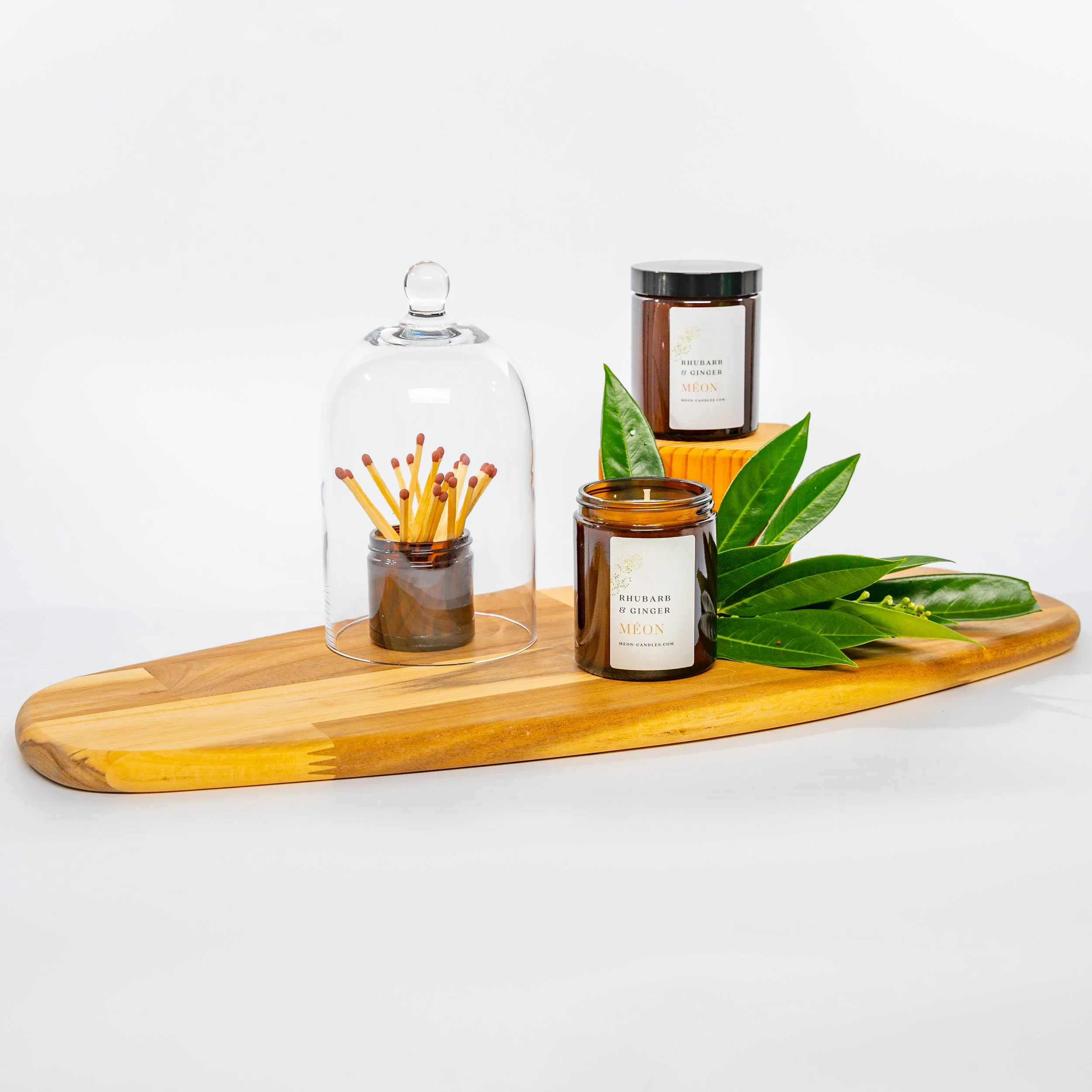 Wooden tray with green leaves, two amber jars labeled 'Rhubarb & Ginger' and candles, a glass covering over a small container of matchsticks.