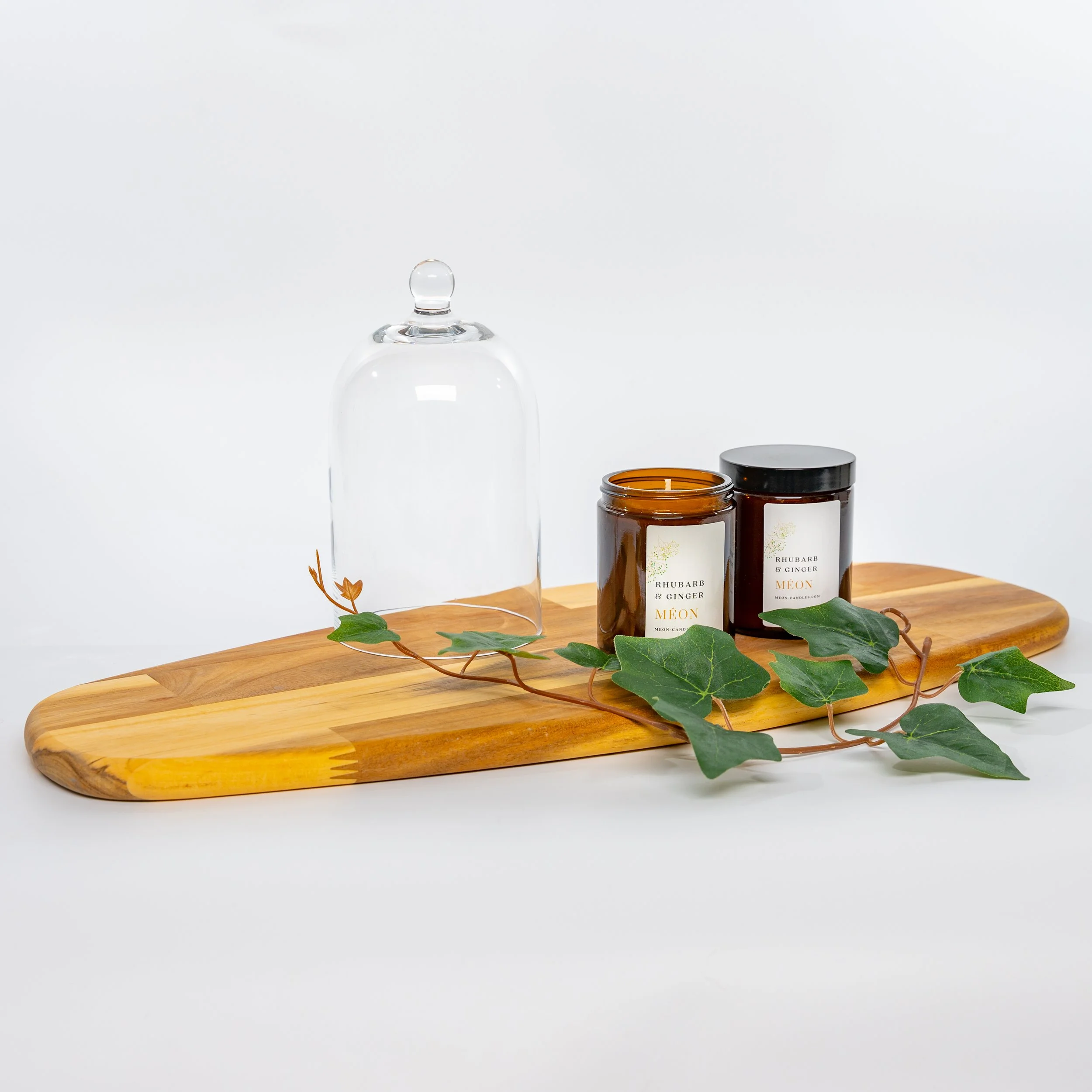 Decorative wooden tray with two candles labeled "Rhubarb & Ginger" and an empty glass cloche, accented with green ivy leaves, on a white background.