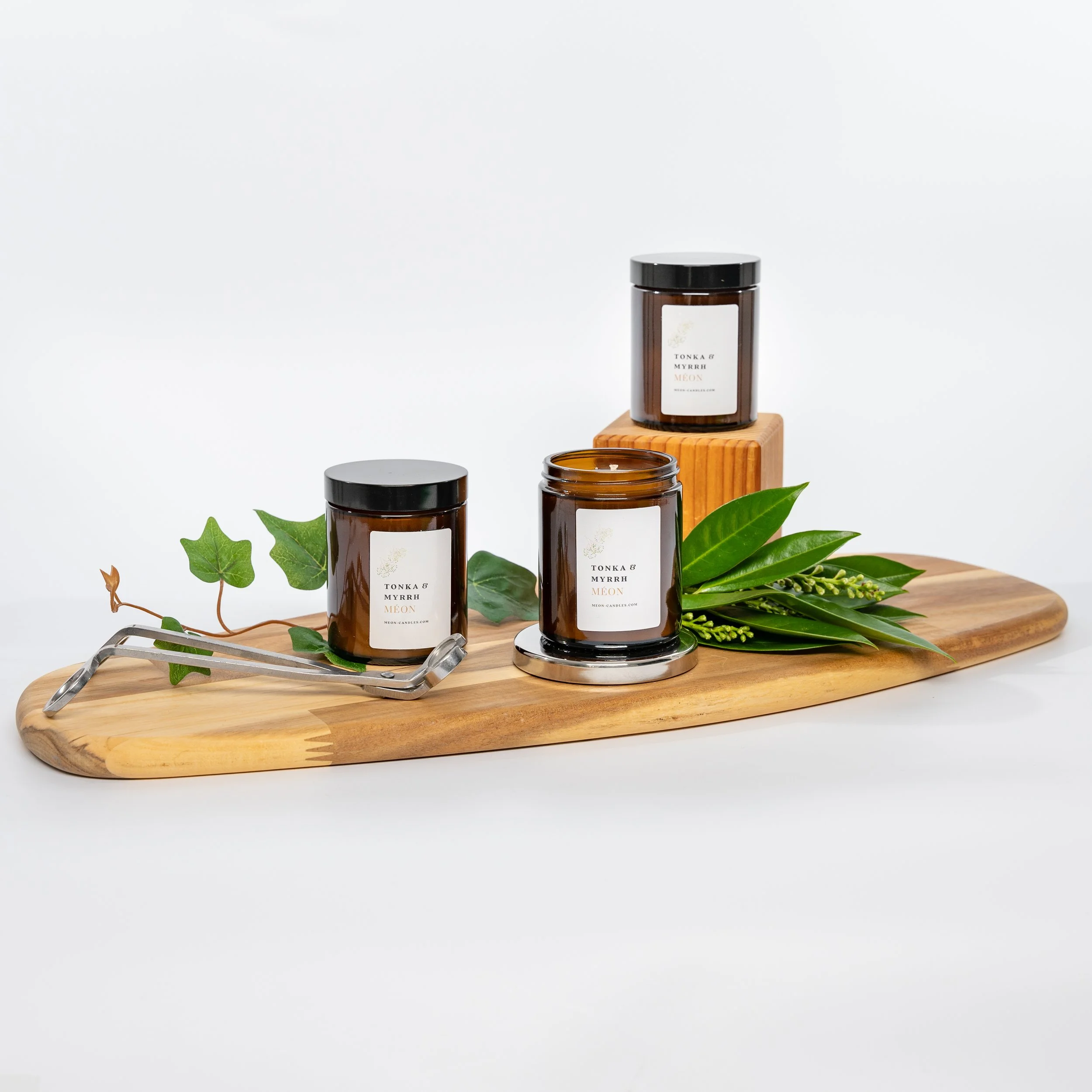 Three amber glass candle jars with black lids, arranged on a wooden board with green leaves and a candle wick snuffer.