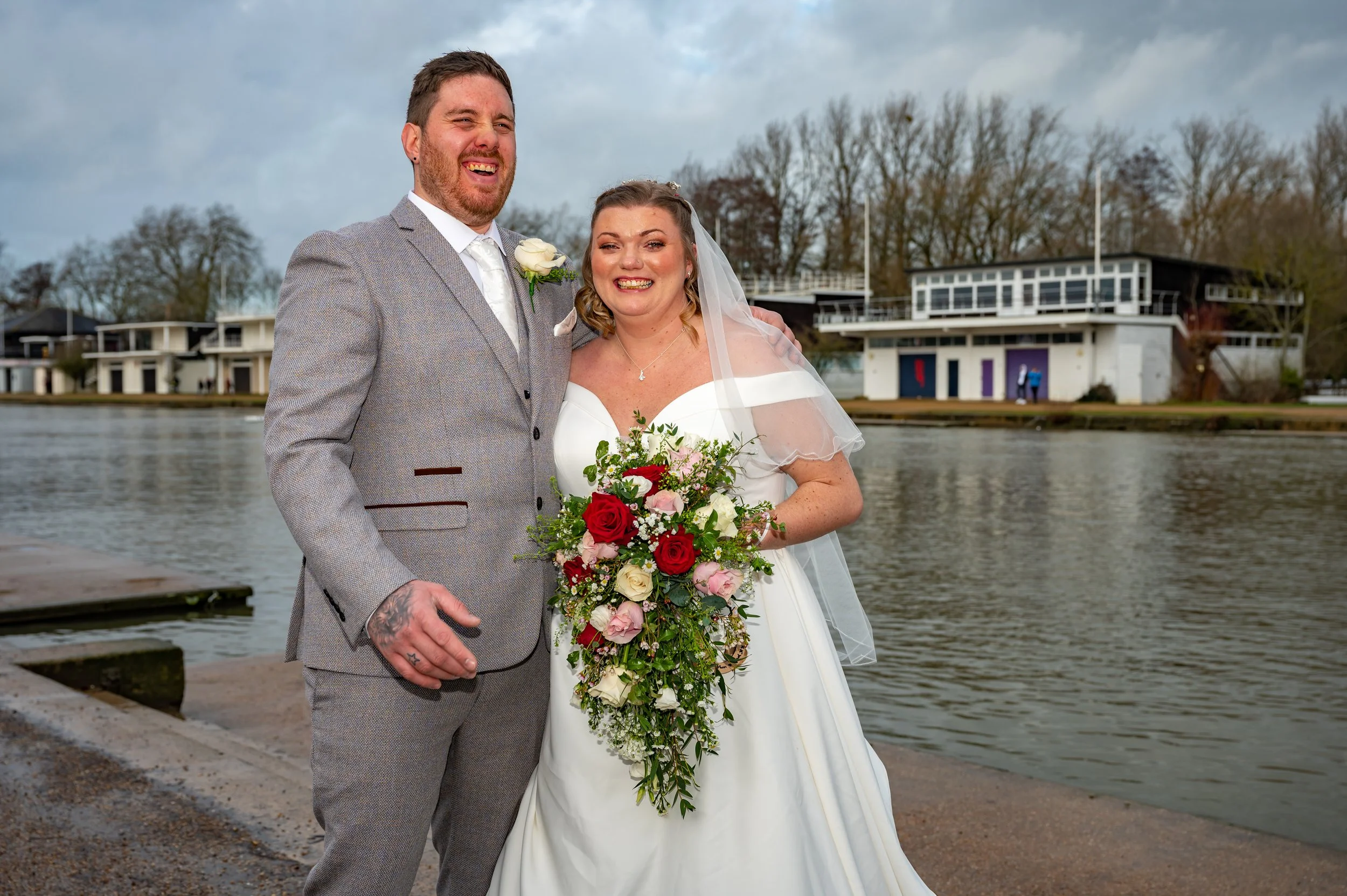 A bride holding her bouquet of flowers stands with her husband next to the River Thames Oxford