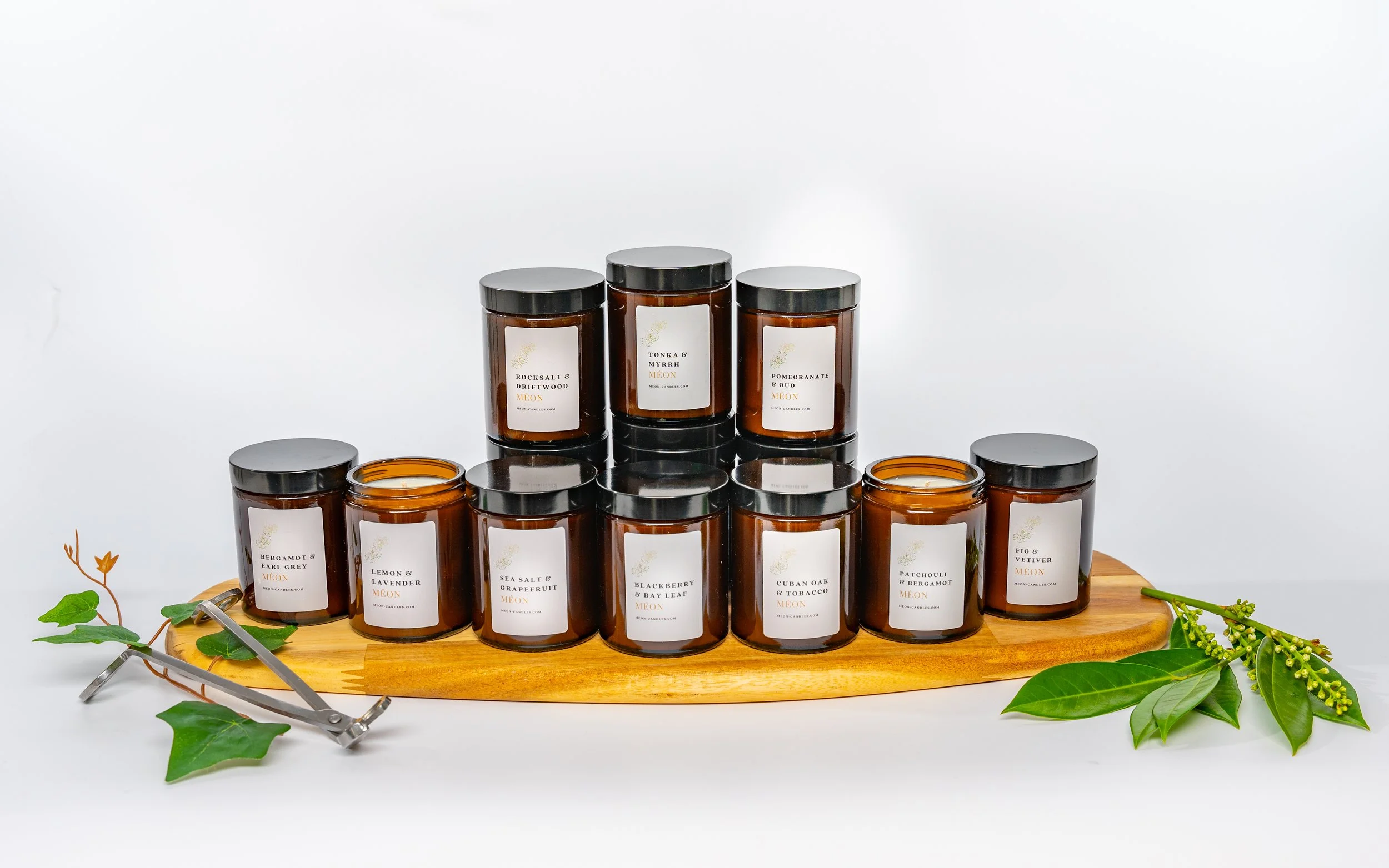 Collection of brown candles in black jars with white labels, arranged on a wooden tray surrounded by green leaves and sprigs of foliage, with a white background.