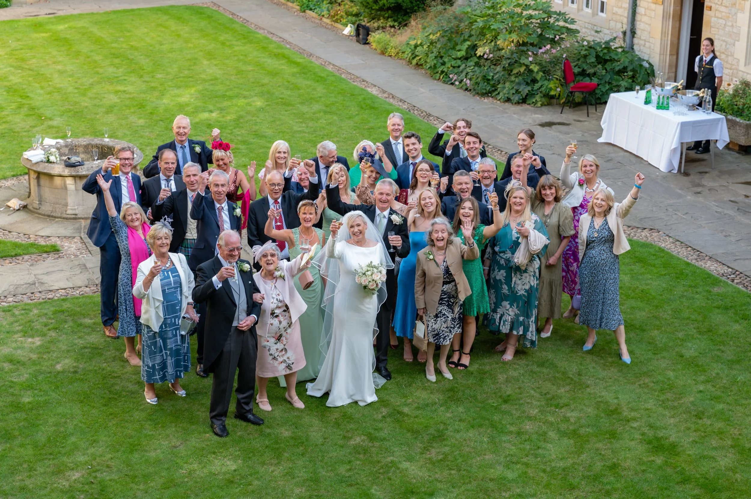 A group of wedding guests stood as a group on grass hold their champagne glasses up in the air